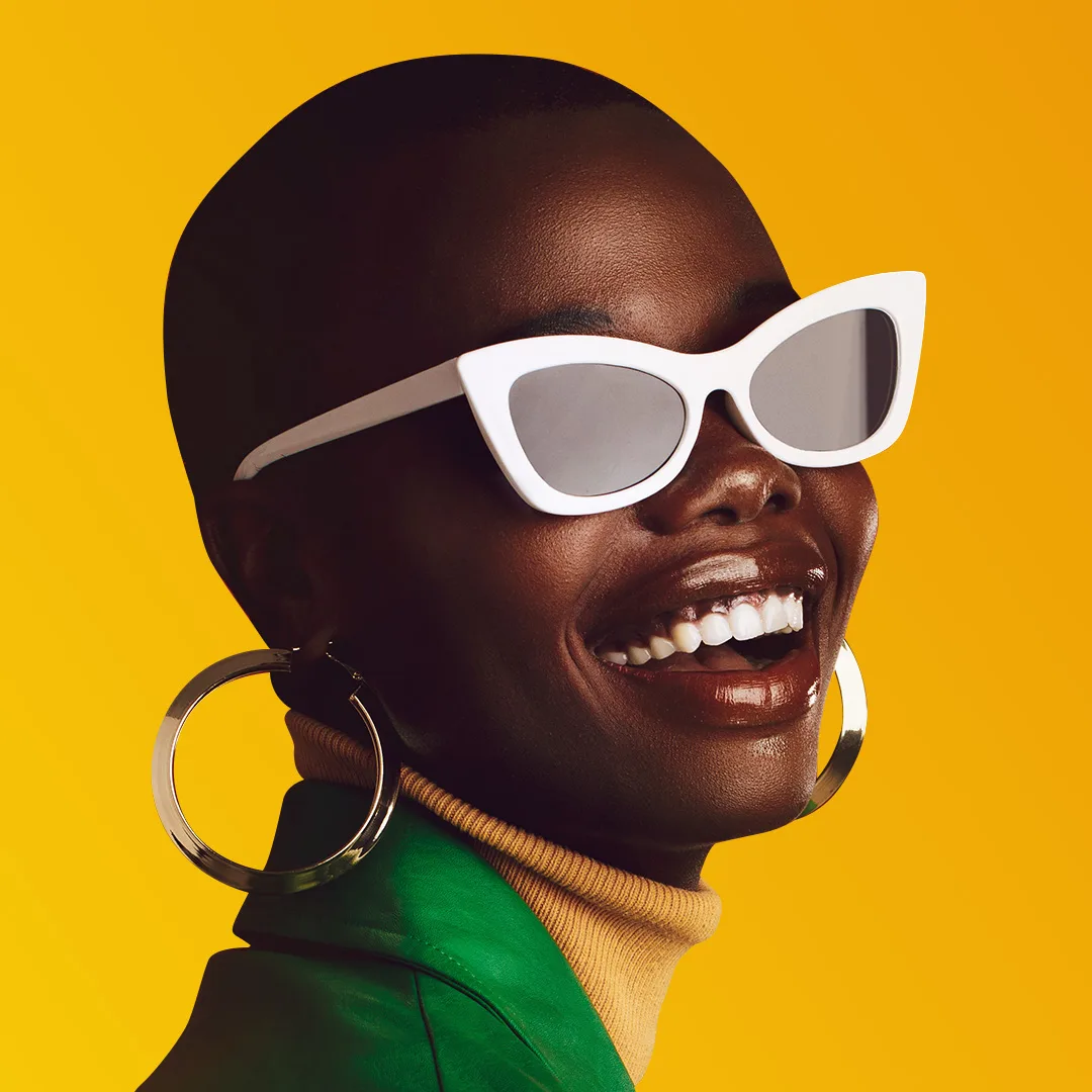 A smiling woman wearing large earrings and white sunglasses against a bright yellow background.