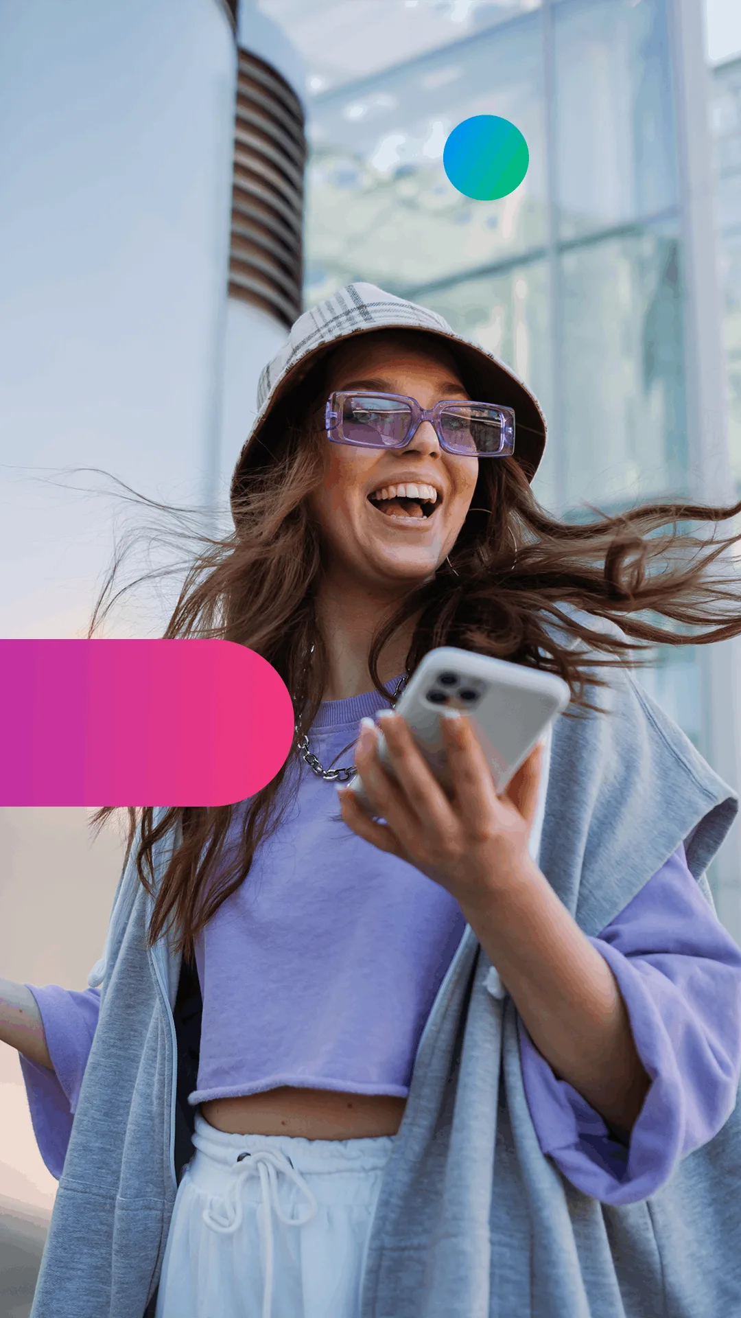 A smiling woman wearing sunglasses and a bucket hat holds a smartphone, with her hair flowing in the wind outdoors.