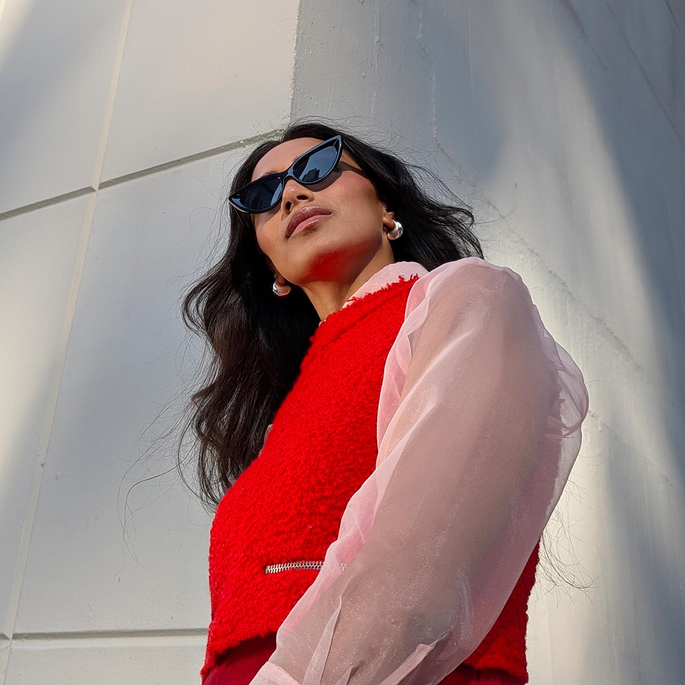 A high-angle fashion portrait of a woman looking upward, wearing black cat-eye sunglasses and a red textured vest with sheer pink sleeves. The shot highlights the camera's ability to capture sharp details and vibrant colors in bright, direct daylight.