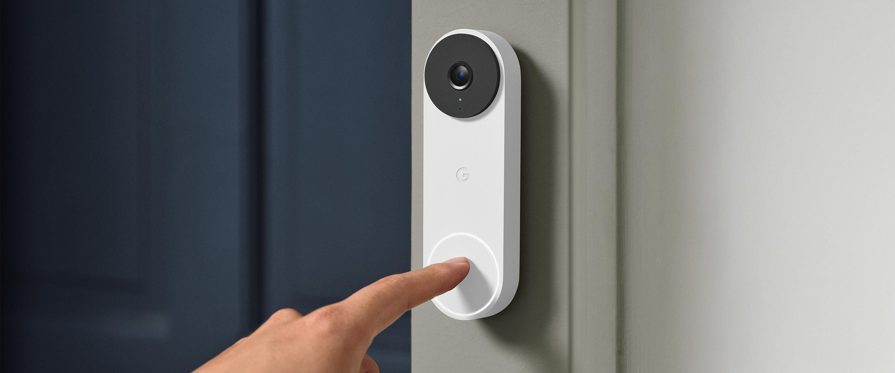 A person's index finger presses the button on a white Google Nest video doorbell mounted on a gray wall next to a dark blue door.