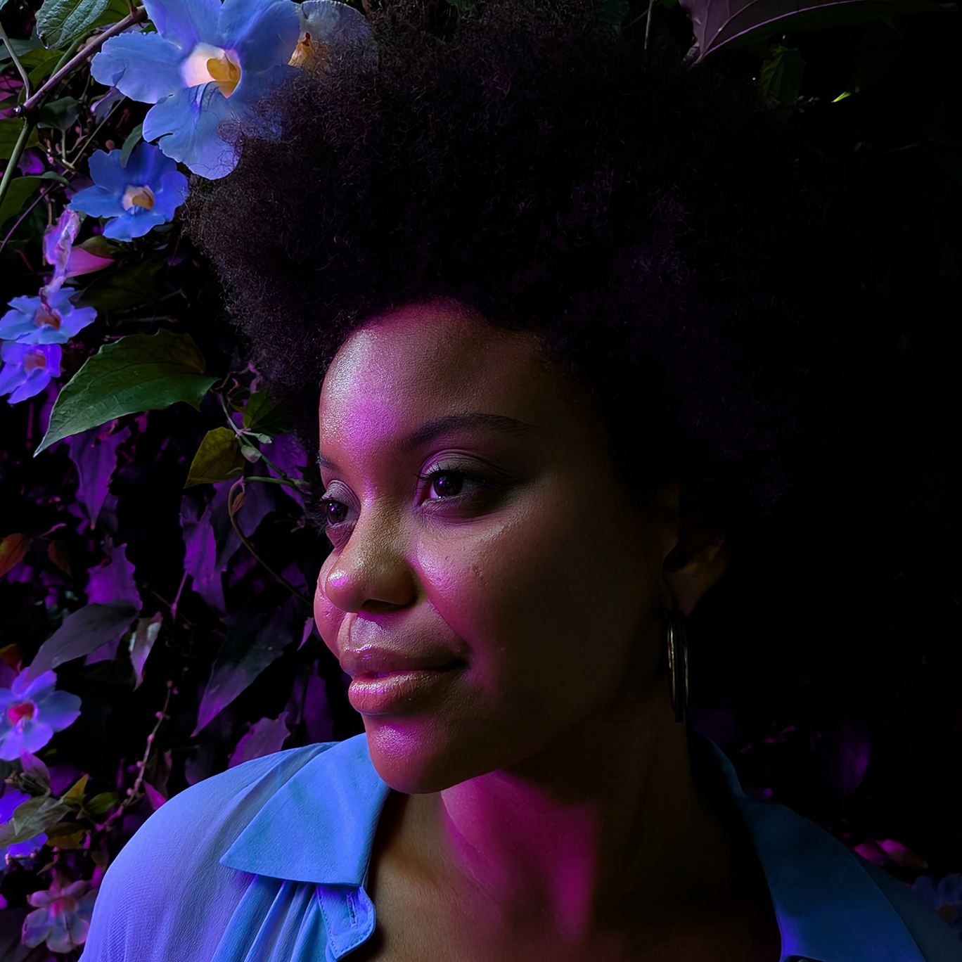 A portrait of a woman with an afro looking to the side, illuminated by vibrant purple and blue ambient light. She is surrounded by blue flowers in a dark, natural setting. The image demonstrates the device's ability to capture rich detail and skin tones in low-light, high-contrast environments.