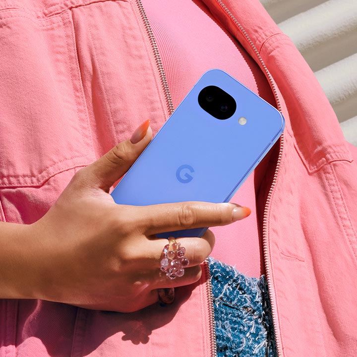 A close-up lifestyle shot of a person’s hand holding a Pixel 10a. The phone is held against a textured pink denim jacket, showcasing the device's sleek, flush-back design where the dual-camera lenses are set directly into the body rather than a raised bar.