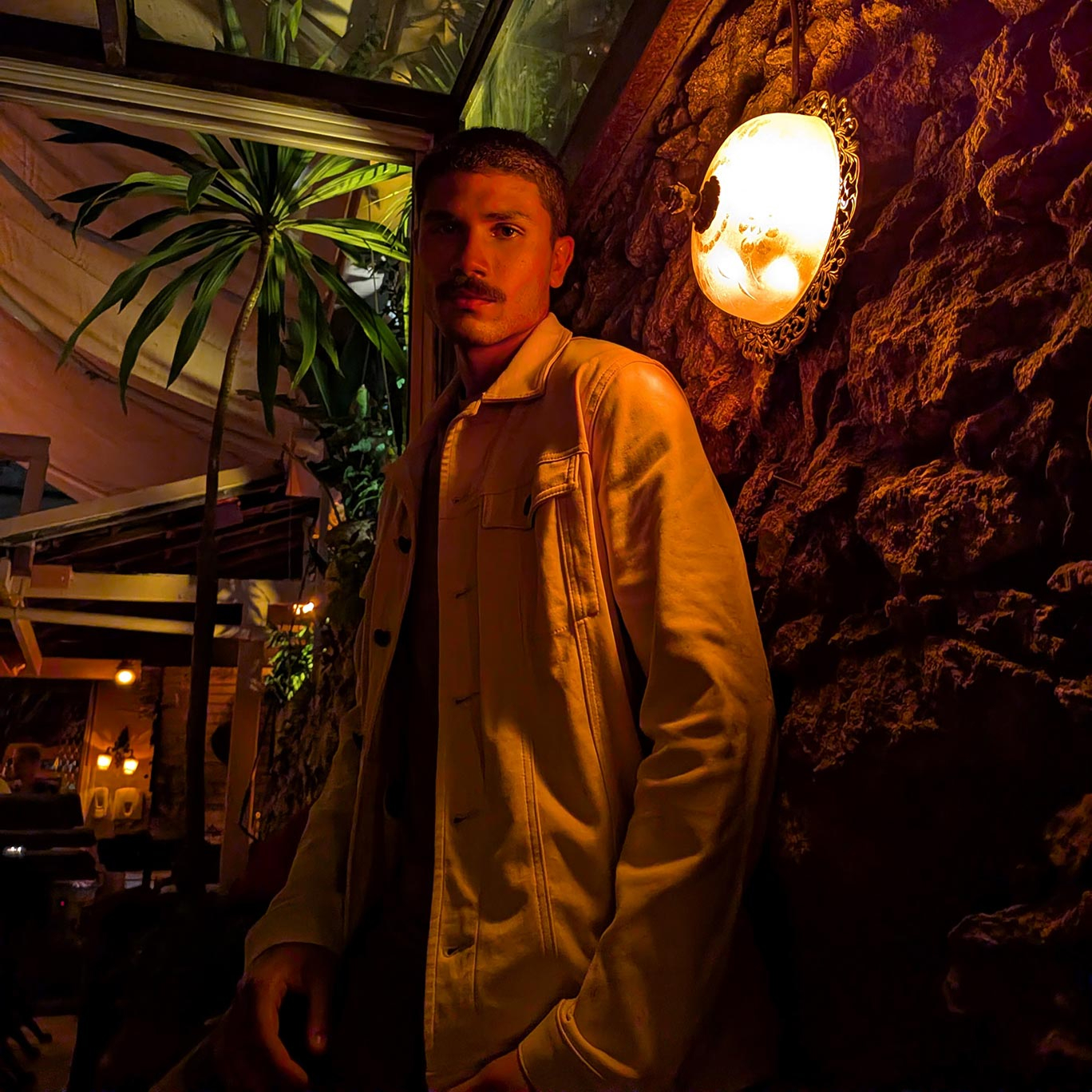 A portrait of a man with a mustache wearing a tan jacket, leaning against a textured stone wall indoors. The image demonstrates the camera's performance in capturing rich textures and warm skin tones in dim, atmospheric indoor lighting.