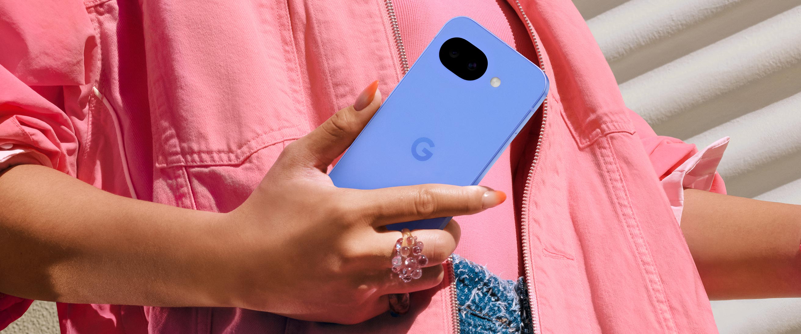 A close-up lifestyle shot of a person’s hand holding a Pixel 10a. The phone is held against a textured pink denim jacket, showcasing the device's sleek, flush-back design where the dual-camera lenses are set directly into the body rather than a raised bar.