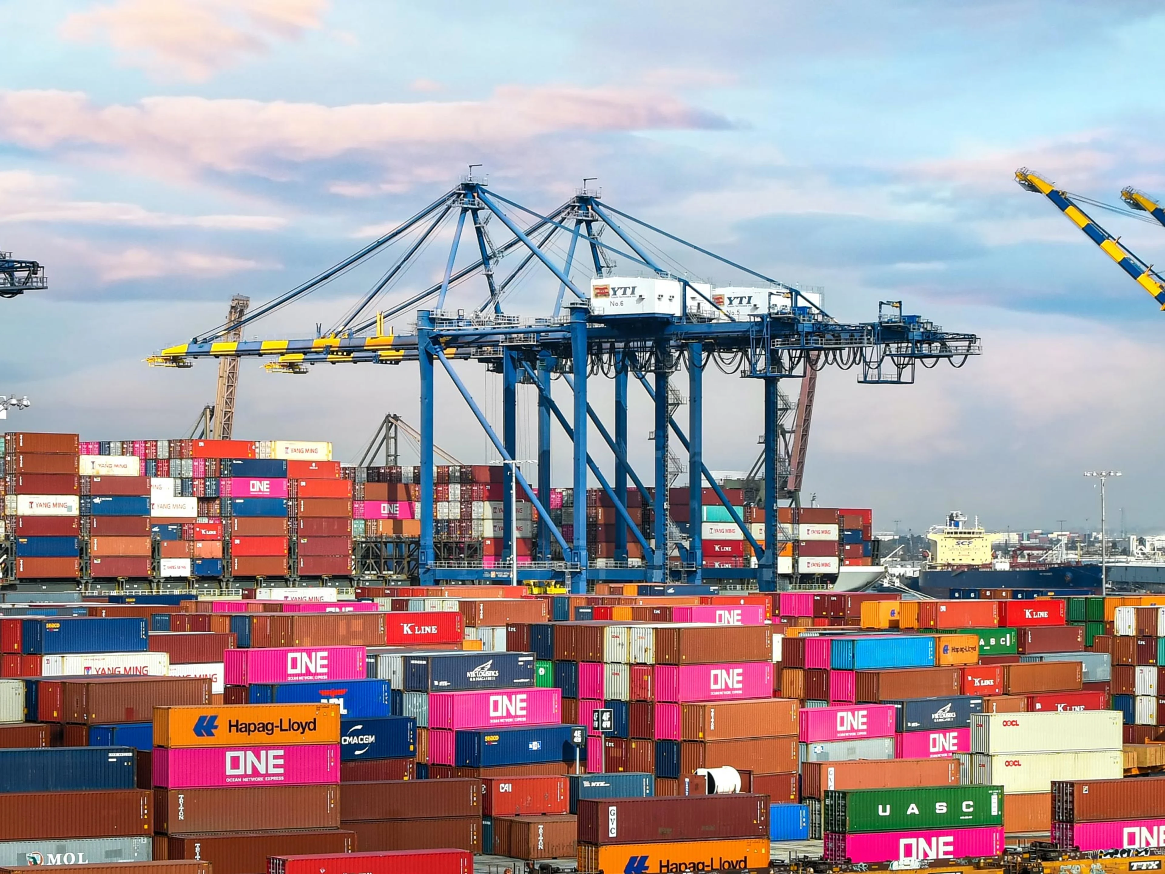 Stacks of colorful shipping containers sit in front of large blue cranes at a busy seaport, with ships and cranes visible in the background under a partly cloudy sky.