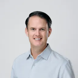 A man with short dark hair wearing a light blue, long-sleeve, button-up shirt smiles at the camera against a plain, light background.