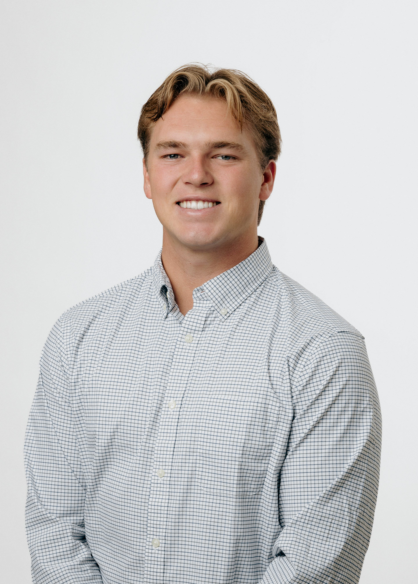 A young man with short, light brown hair smiles at the camera. He is wearing a light-colored, checkered button-up shirt and is posed against a plain white background.