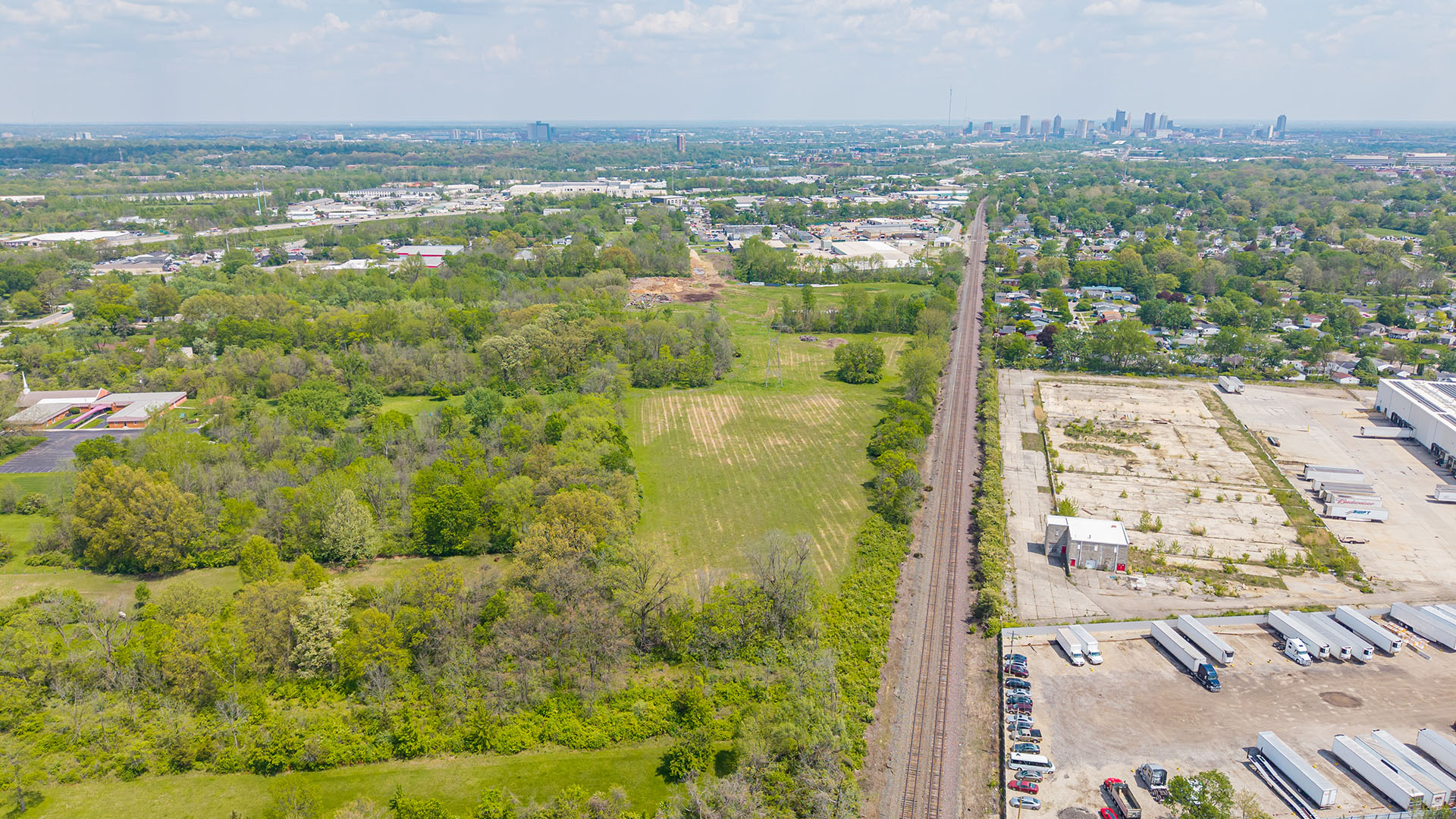 Aerial view of a railway line running alongside green fields, trees, industrial buildings, and parked trucks, with a distant city skyline under a partly cloudy sky.