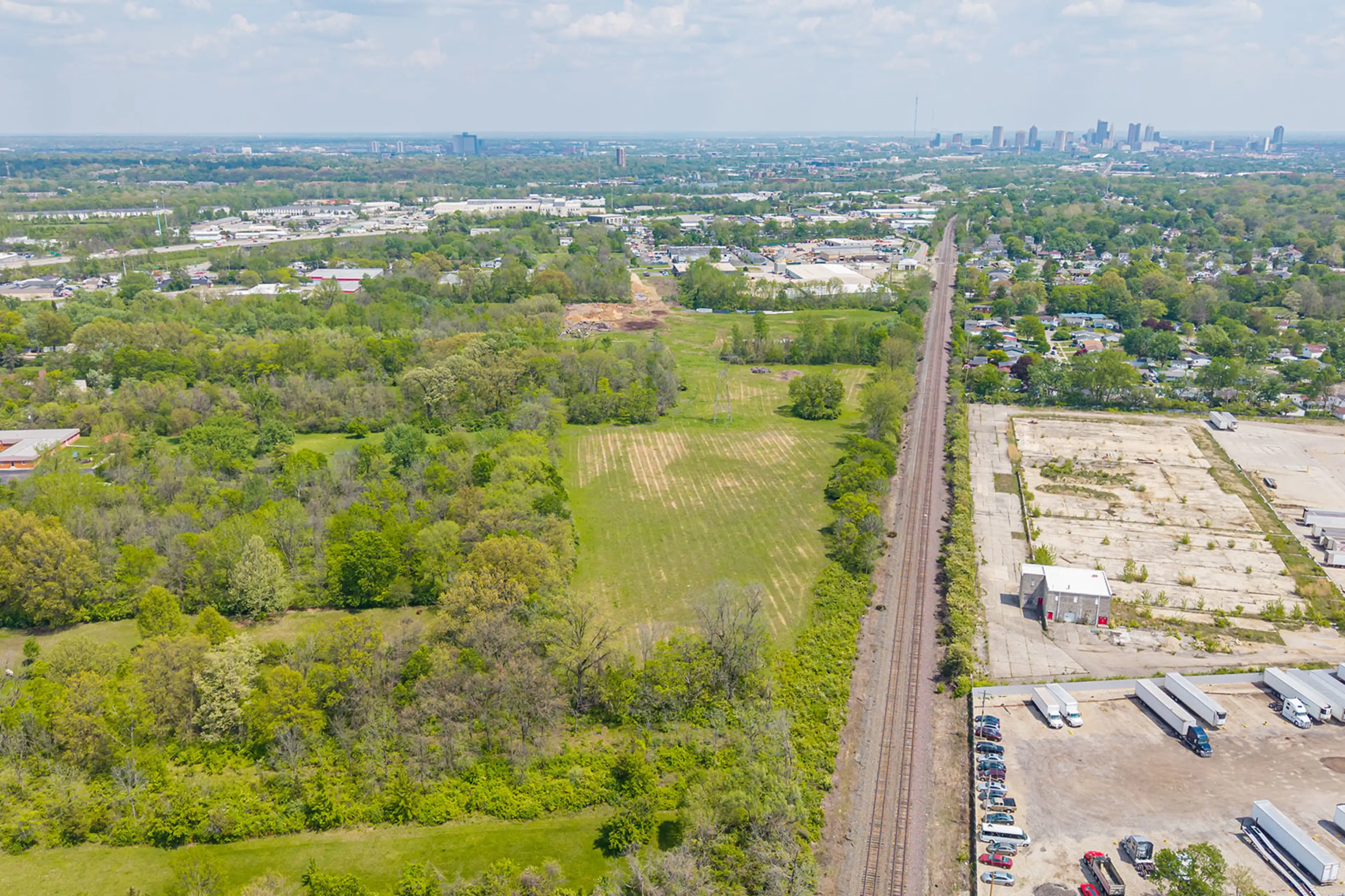 Aerial view of a railway line running alongside green fields, trees, industrial buildings, and parked trucks, with a distant city skyline under a partly cloudy sky.