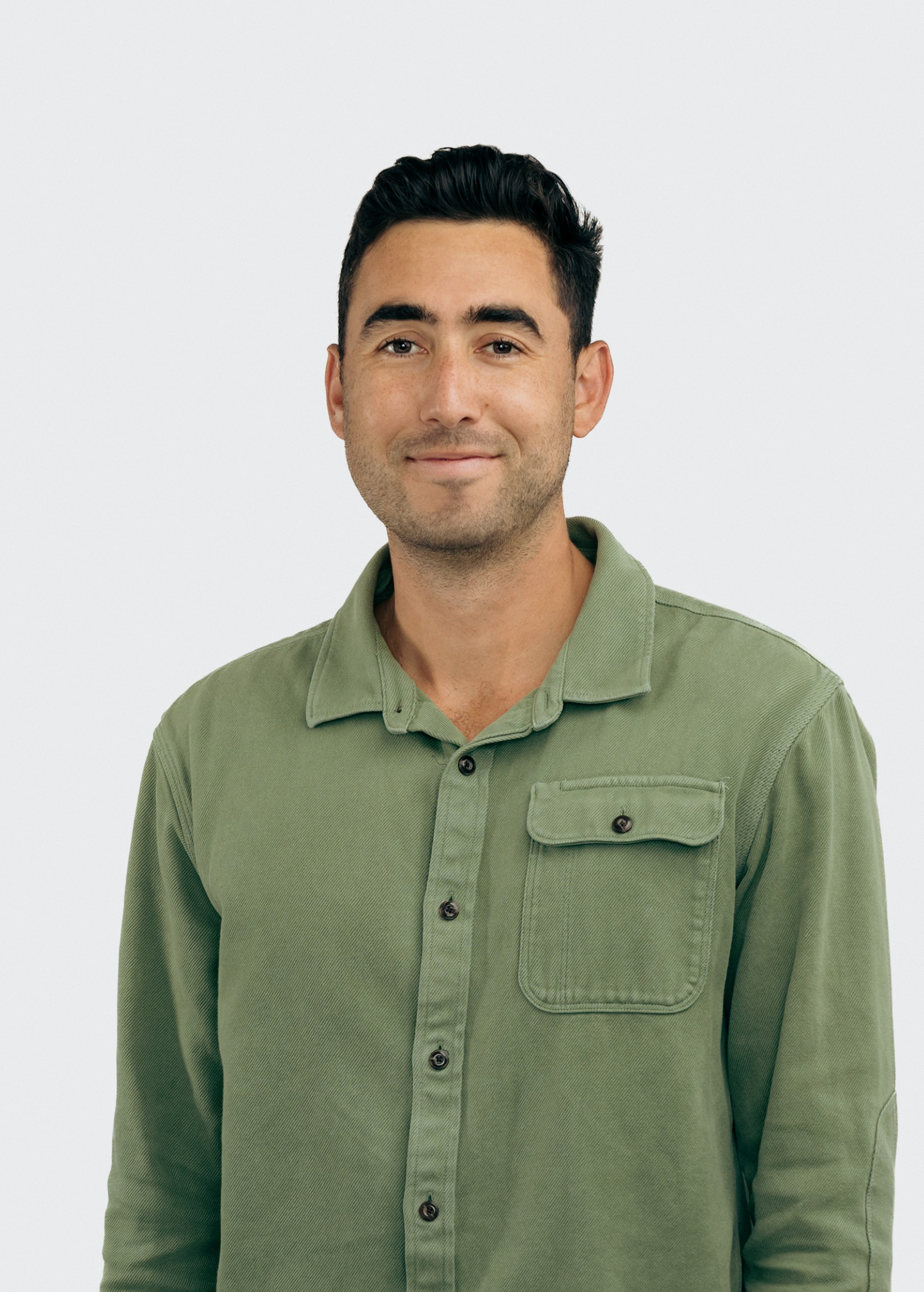 A man with short dark hair wearing a green button-up shirt is smiling softly at the camera against a plain light gray background.
