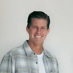 A man with short dark hair is smiling, wearing a light plaid shirt over a white t-shirt, standing against a plain light background.