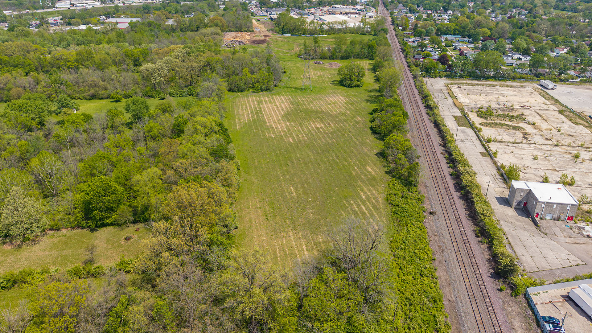 Aerial view of a grassy field bordered by trees, with a set of railroad tracks running alongside an industrial area on the right, and a residential neighborhood visible in the distance.