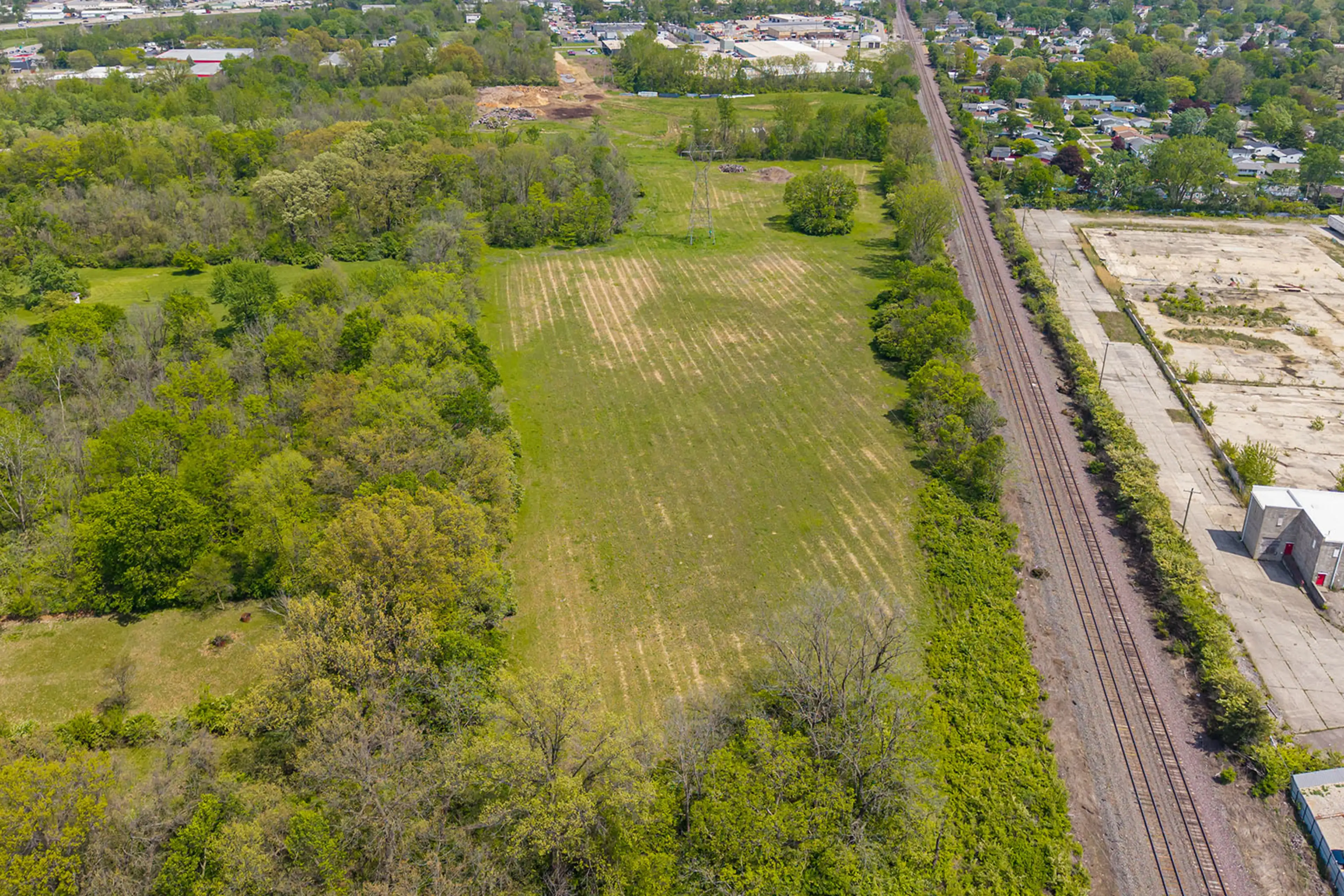 Aerial view of a grassy field bordered by trees, with a set of railroad tracks running alongside an industrial area on the right, and a residential neighborhood visible in the distance.