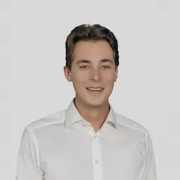 A young man with short brown hair, wearing a white button-up shirt, stands against a plain light gray background, smiling softly at the camera.