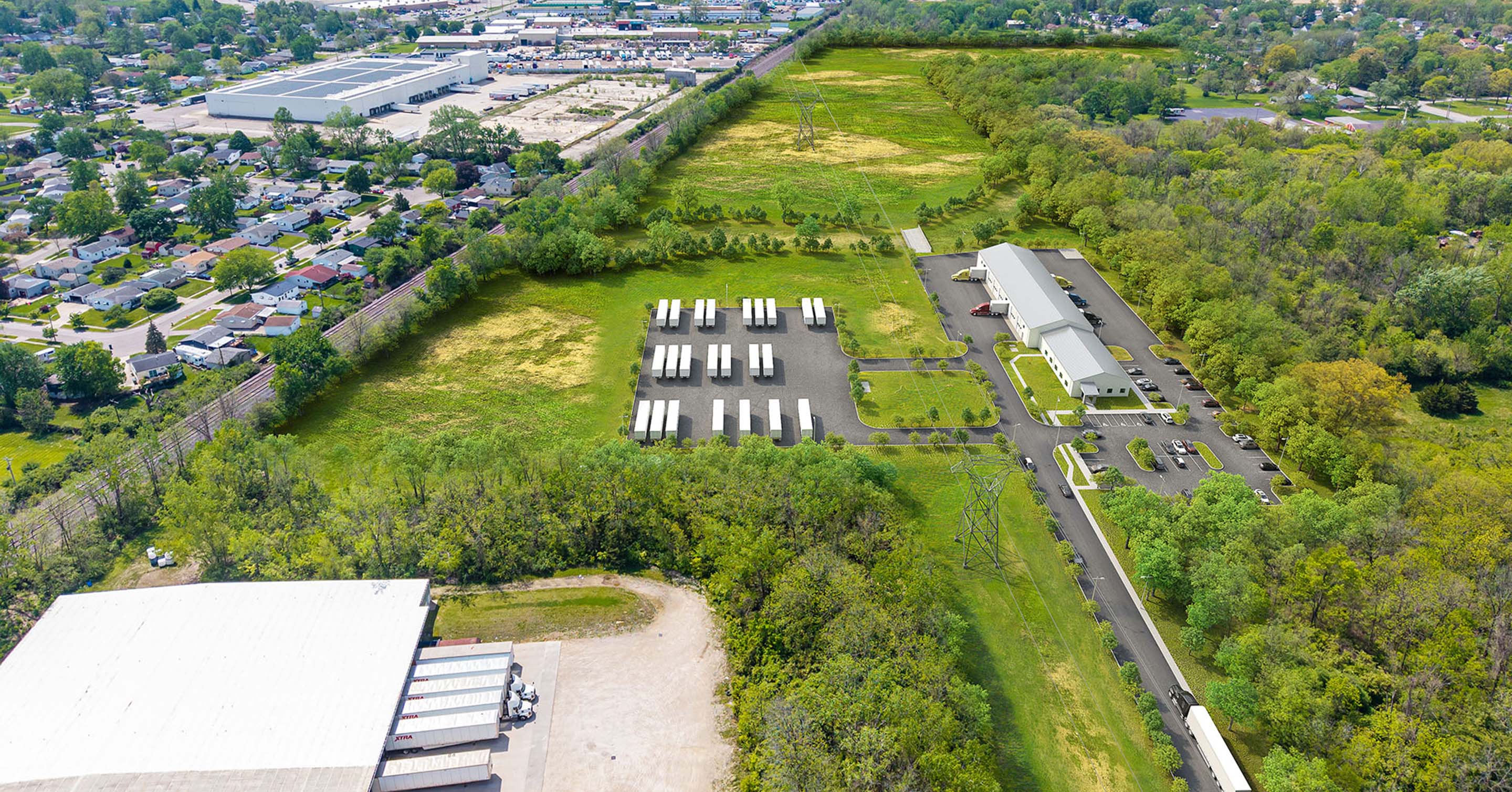 Aerial view of a parking lot with several white trucks, a long building, green fields, trees, and a nearby residential neighborhood.