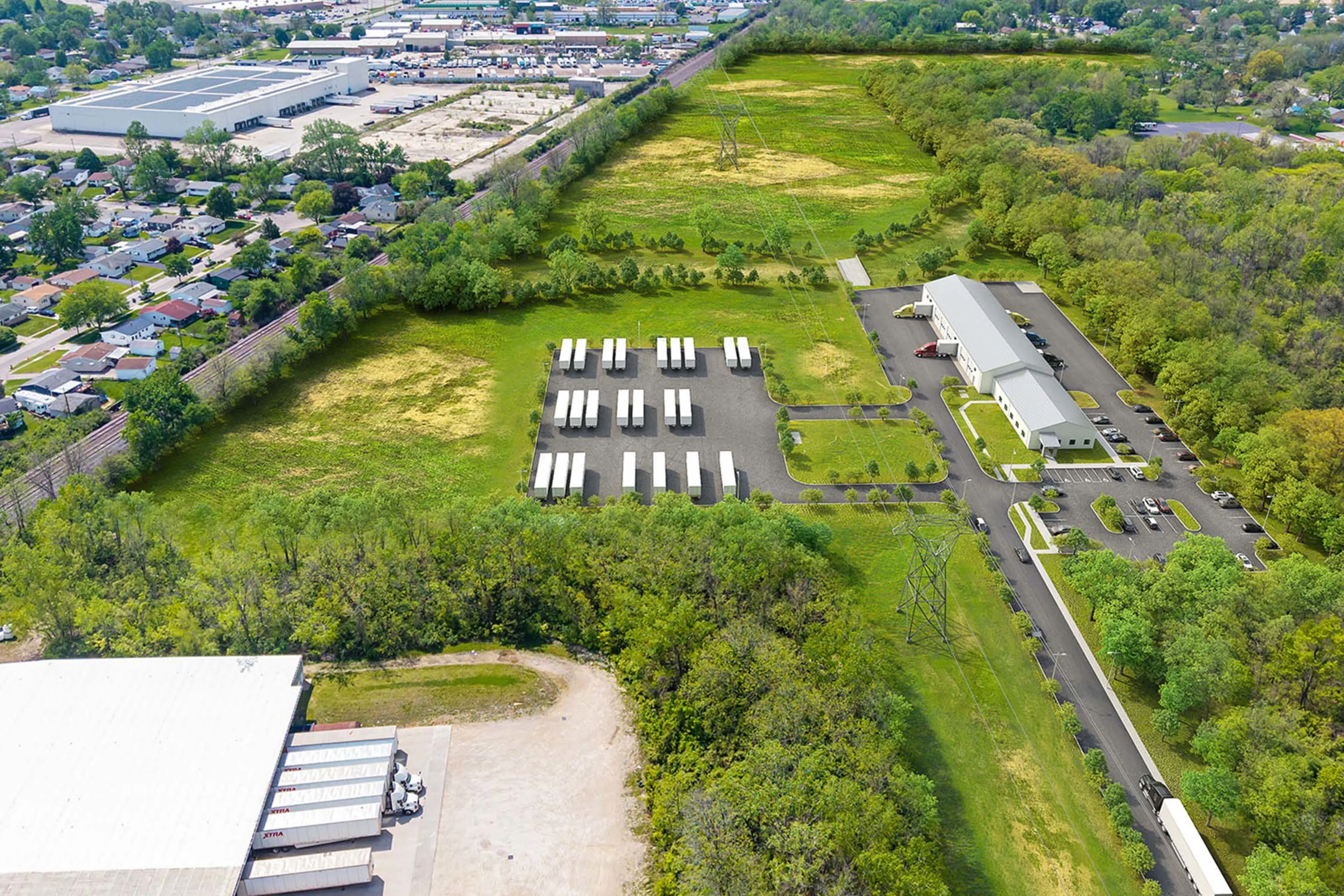 Aerial view of a parking lot with several white trucks, a long building, green fields, trees, and a nearby residential neighborhood.
