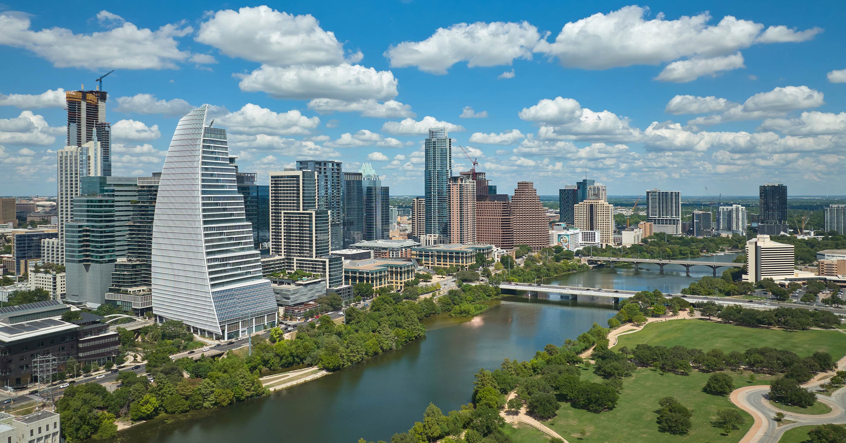 Aerial view of downtown Austin, Texas, featuring modern skyscrapers, a river with bridges, green parks, and a bright blue sky with scattered clouds.