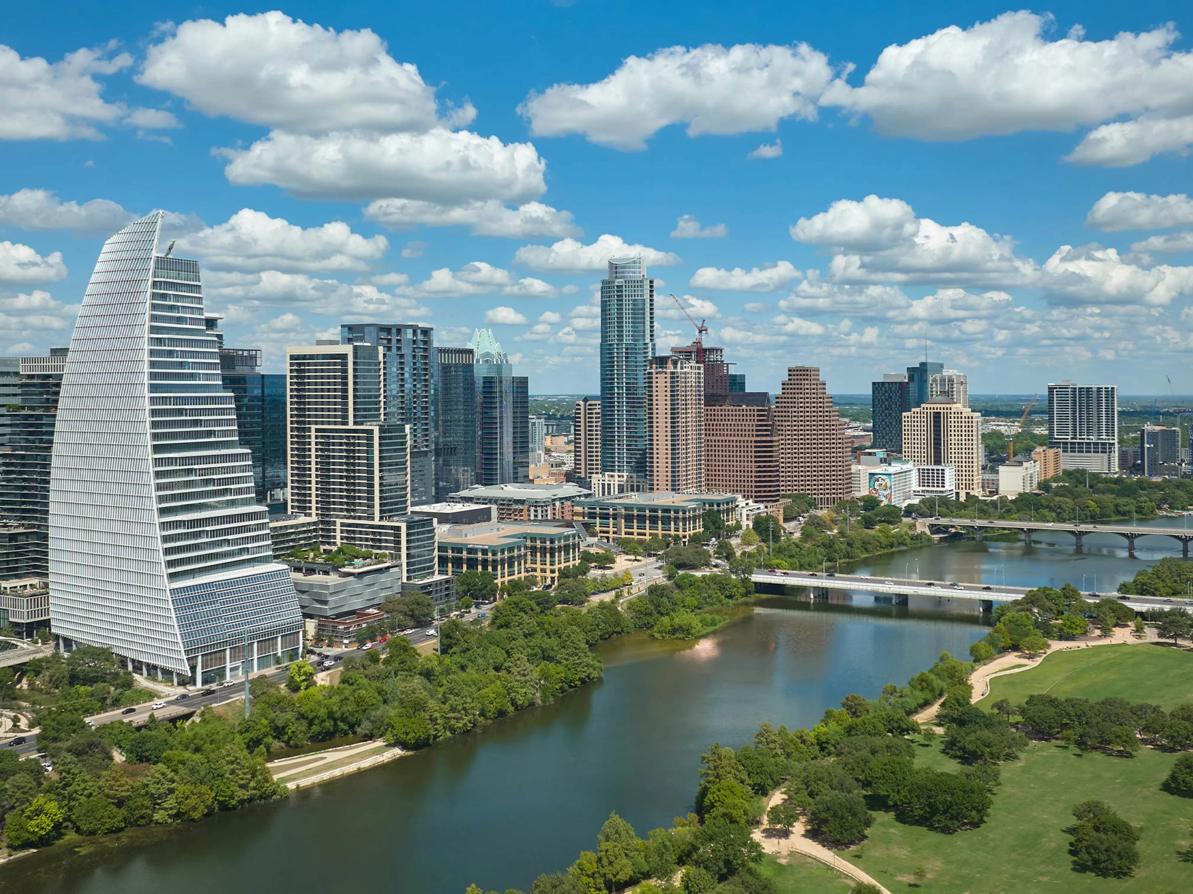 A vibrant city skyline features modern high-rise buildings along a river, with a park and green trees in the foreground under a blue sky with scattered clouds.