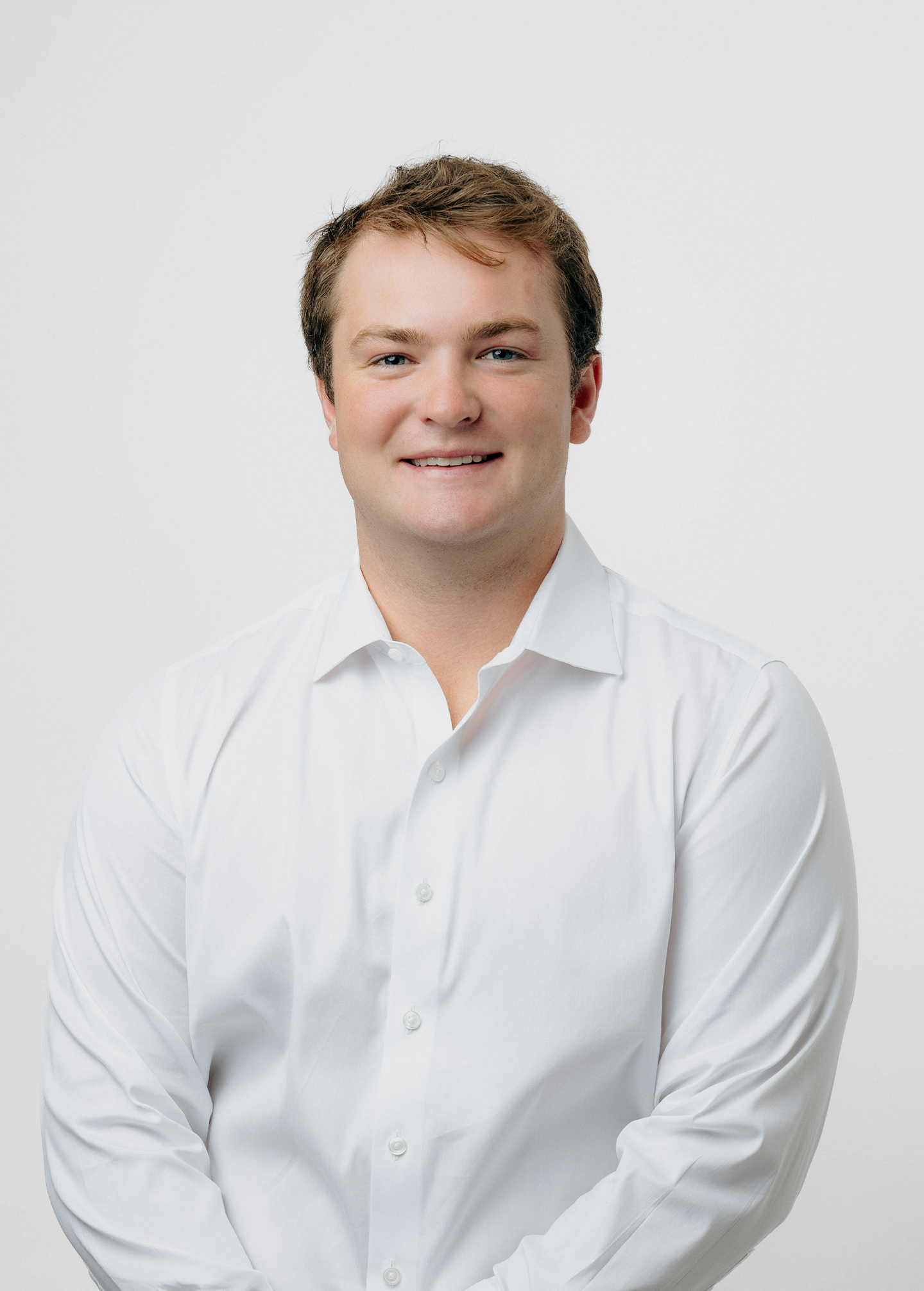 A person with short brown hair, wearing a white button-up shirt, smiles at the camera against a plain white background.
