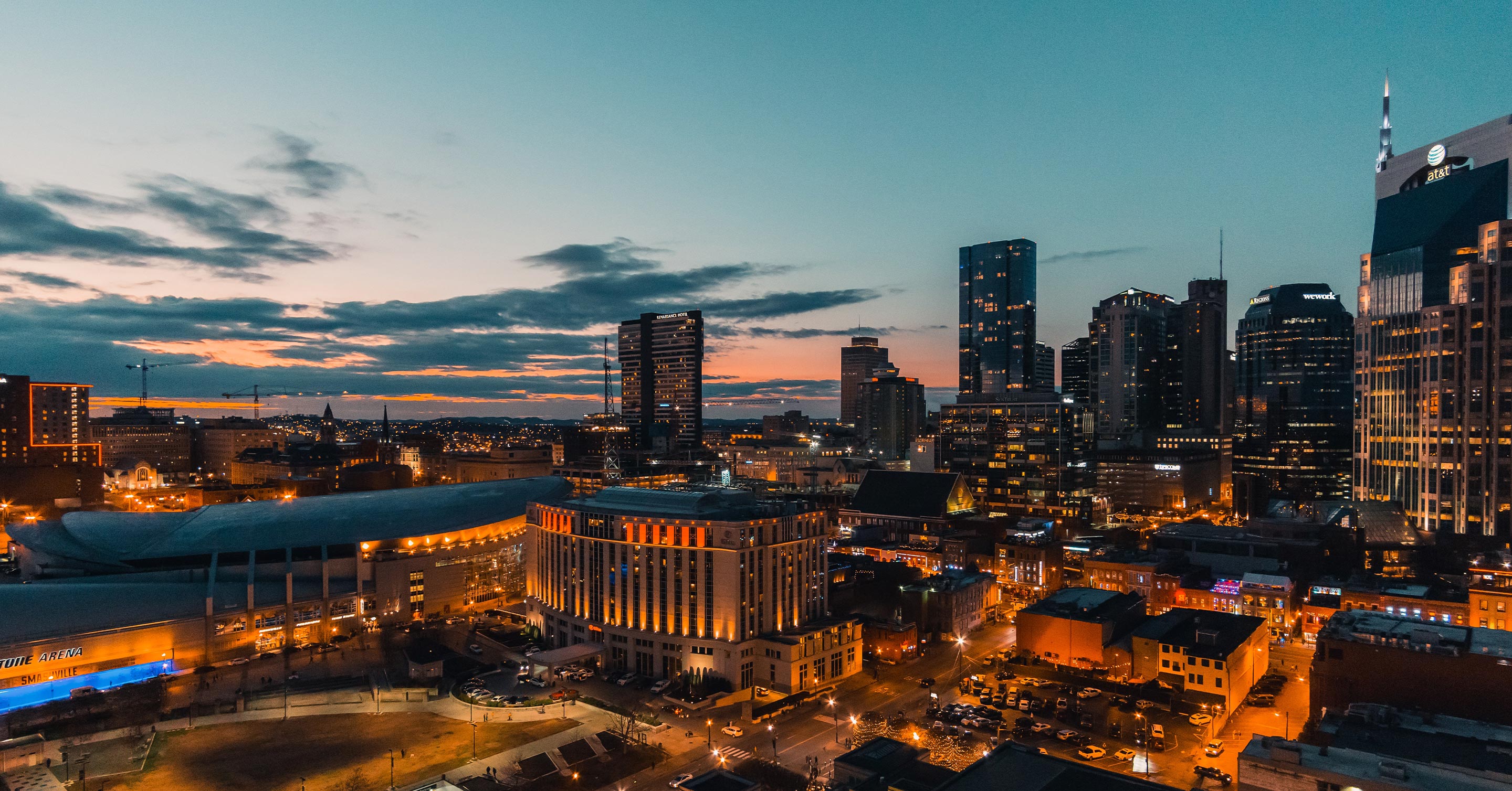 A cityscape at dusk with high-rise buildings illuminated, street lights glowing, and a dramatic sky with clouds and the last light of sunset in the background.