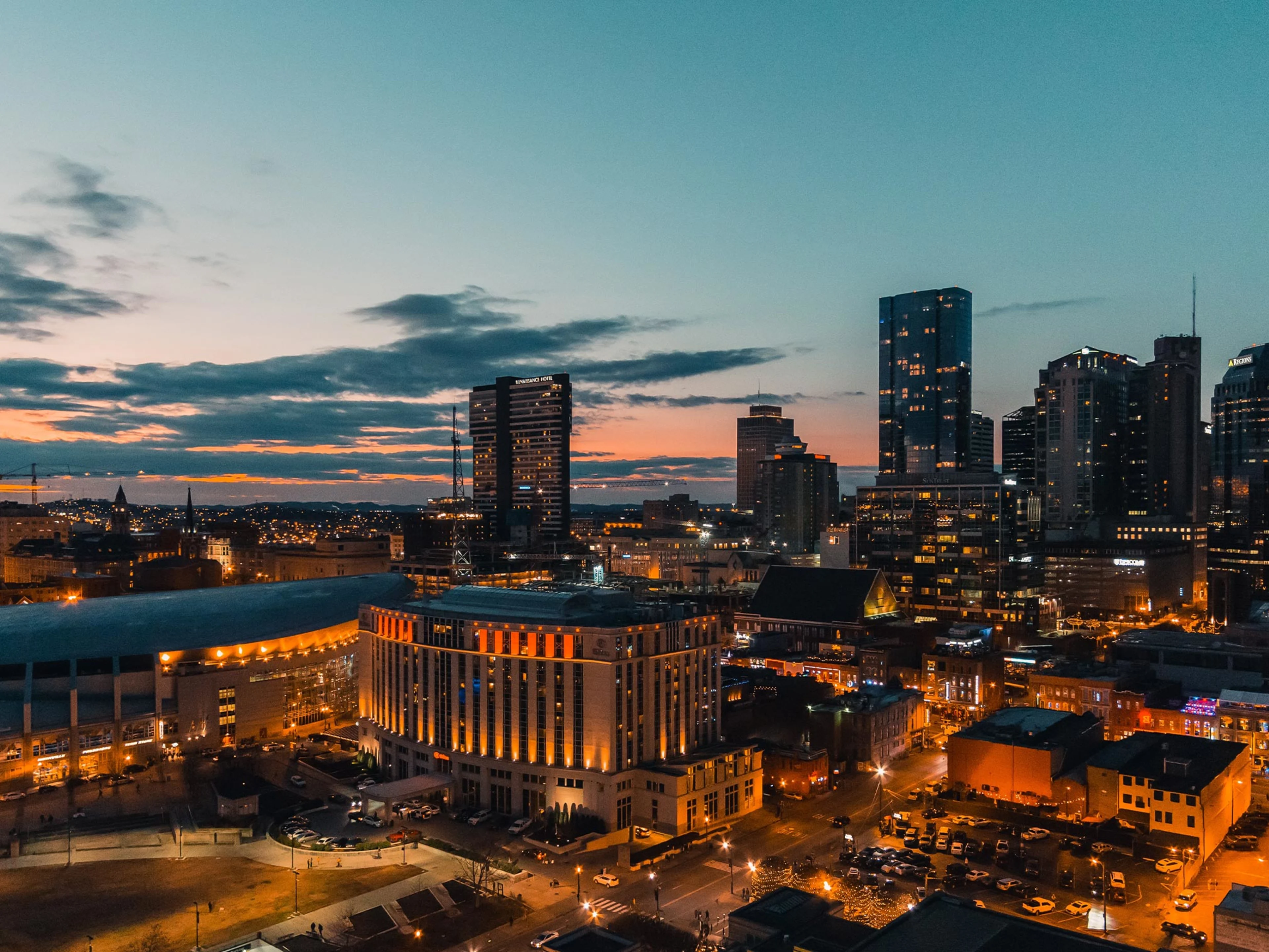 A cityscape at dusk with high-rise buildings illuminated, street lights glowing, and a dramatic sky with clouds and the last light of sunset in the background.
