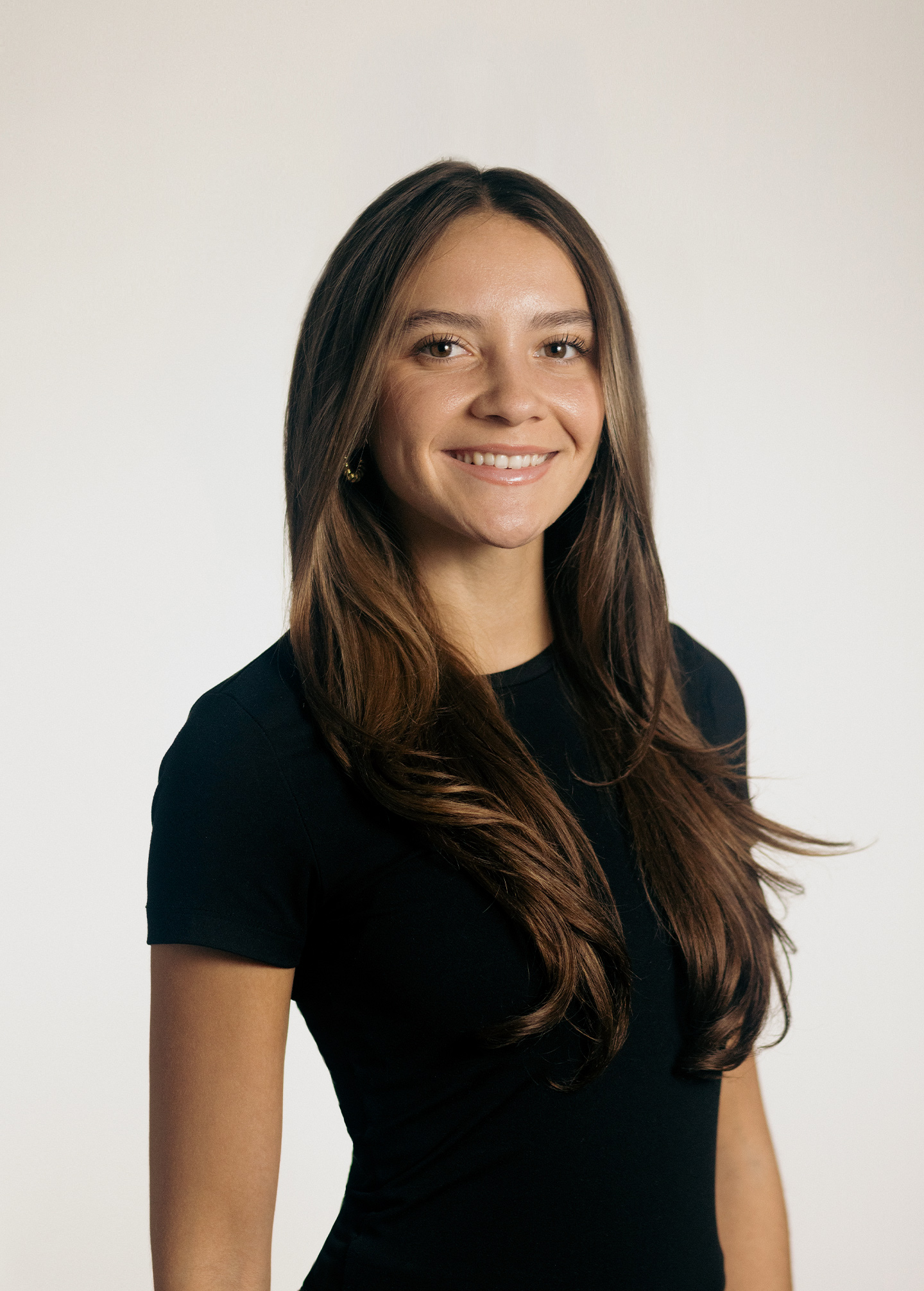 A professional headshot of a young woman with long, layered brown hair, wearing a black t-shirt. She is smiling at the camera against a plain, light gray background.