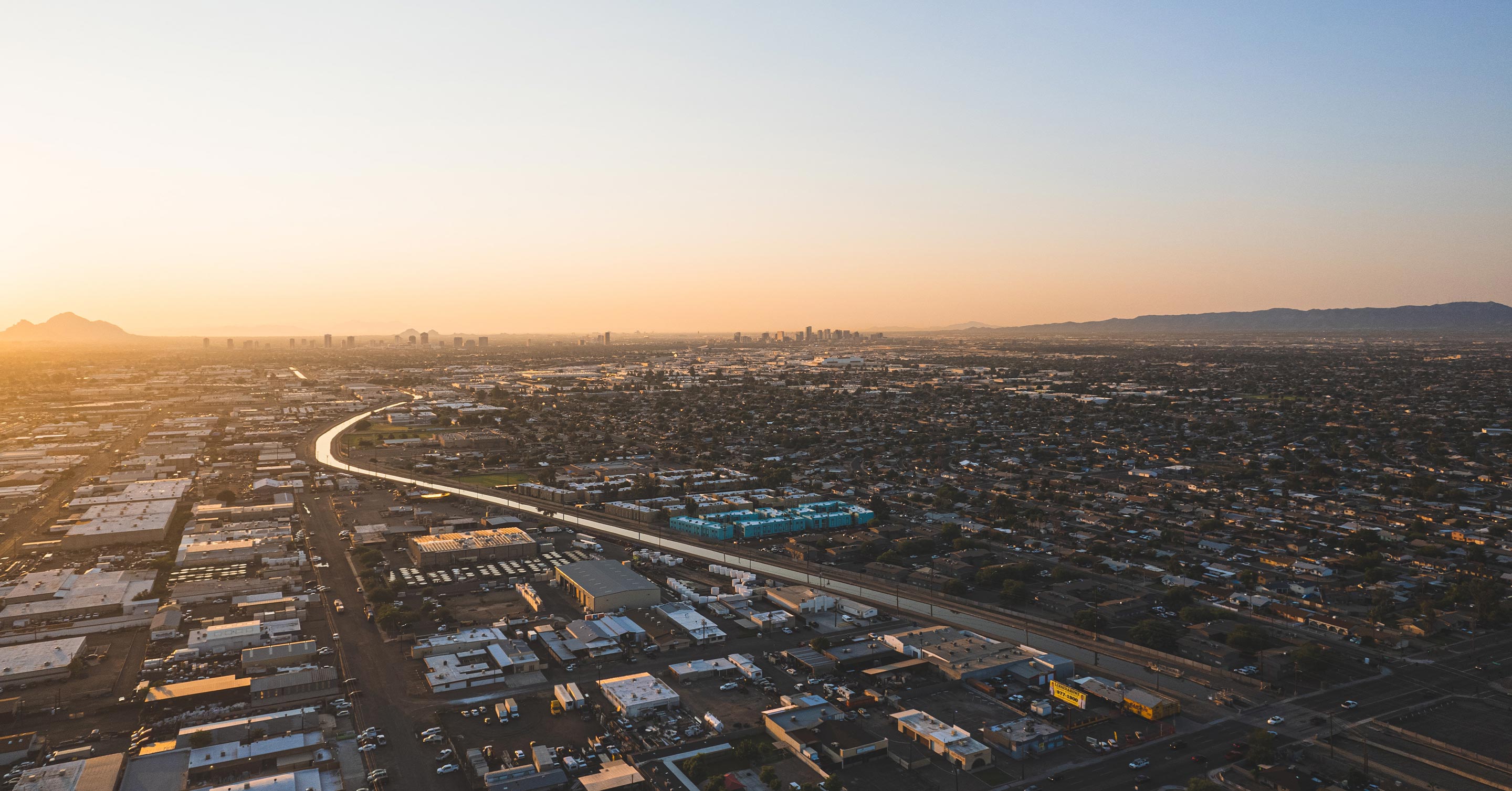 Aerial view of a sprawling cityscape at sunset with a curving road, densely packed buildings, and distant mountains under a clear sky.