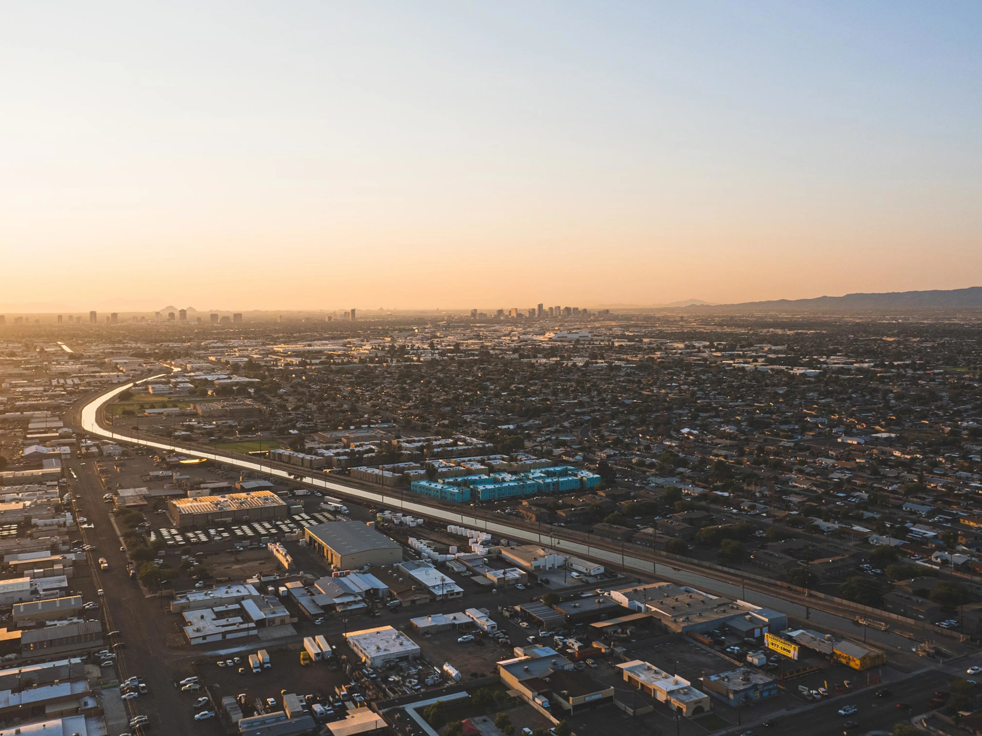 Aerial view of a sprawling cityscape at sunset with a curving road, densely packed buildings, and distant mountains under a clear sky.