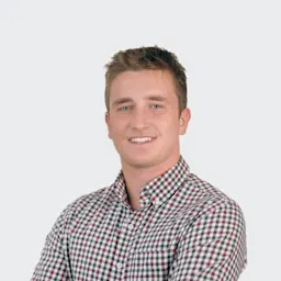 A young man with short brown hair, wearing a checkered button-up shirt, stands with arms crossed and smiles at the camera against a plain light background.