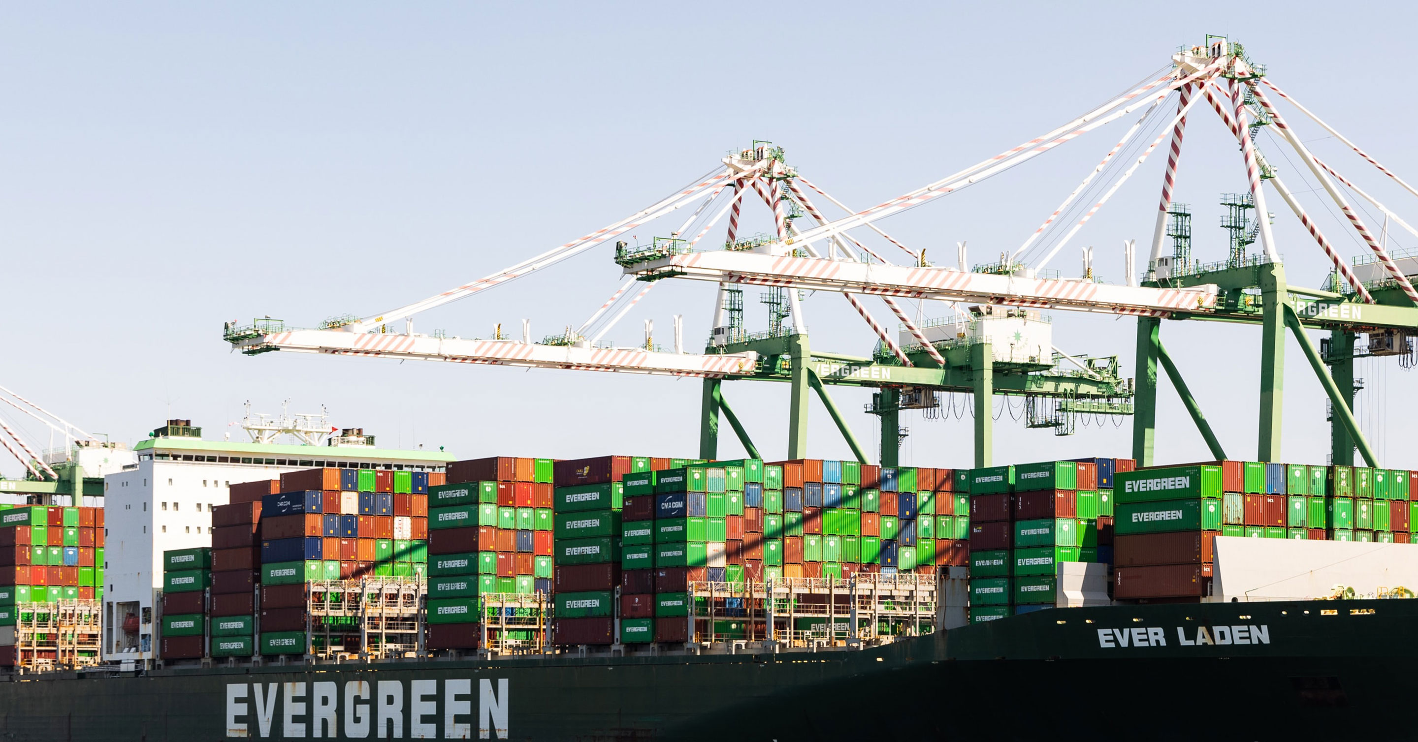A large container ship labeled EVERGREEN is docked at a port, loaded with multicolored shipping containers. Tall cargo cranes tower overhead, ready to move containers on and off the vessel.