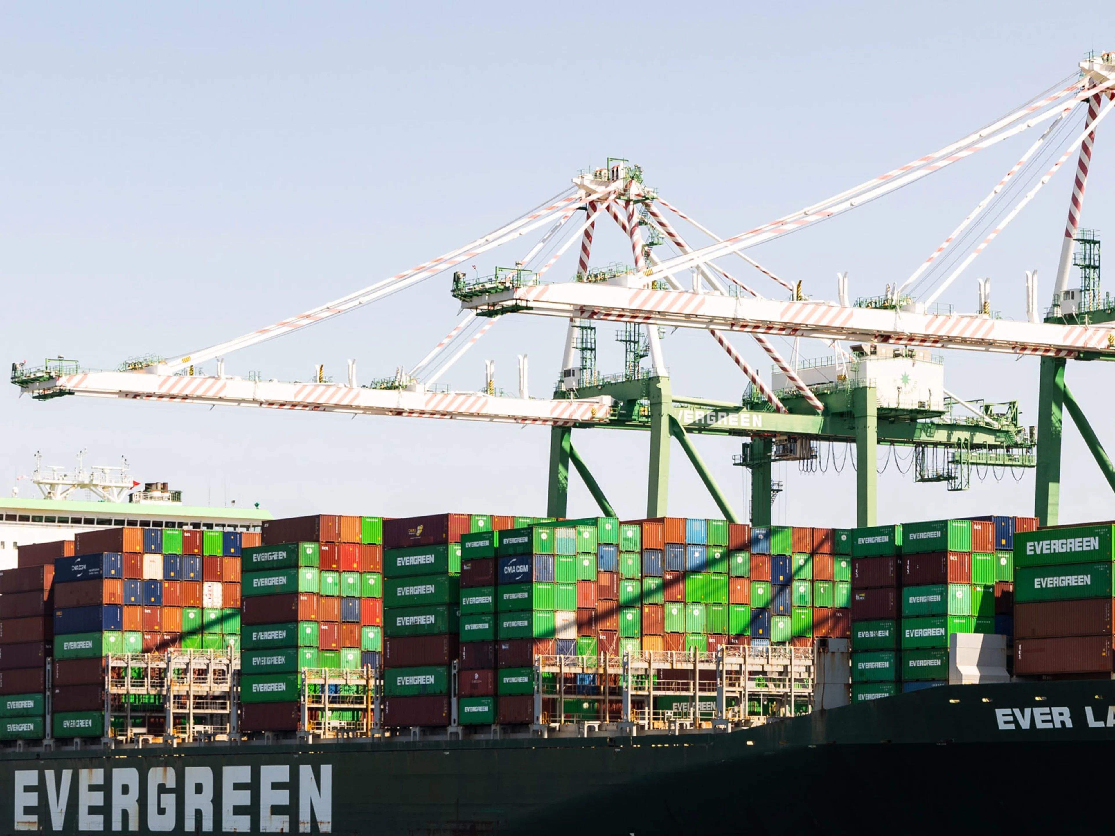 A large container ship labeled EVERGREEN is docked at a port, loaded with multicolored shipping containers. Tall cargo cranes tower overhead, ready to move containers on and off the vessel.