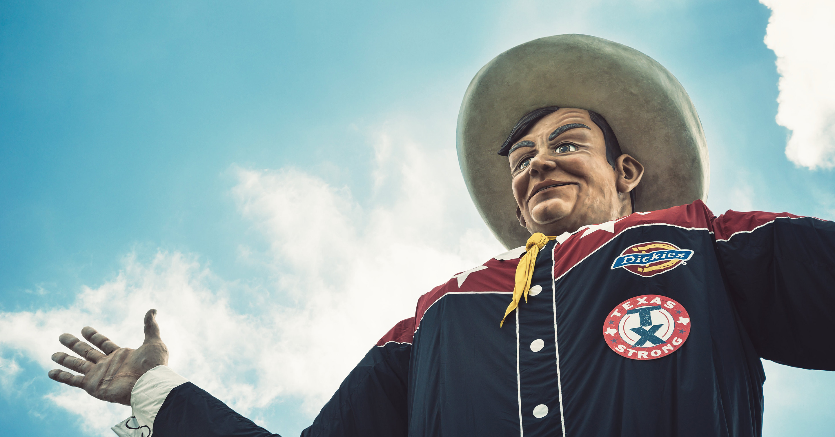 A large cowboy statue in a wide-brimmed hat and dark shirt stands with arms open against a blue sky with clouds. The shirt features patches, including one reading TEXAS STRONG.