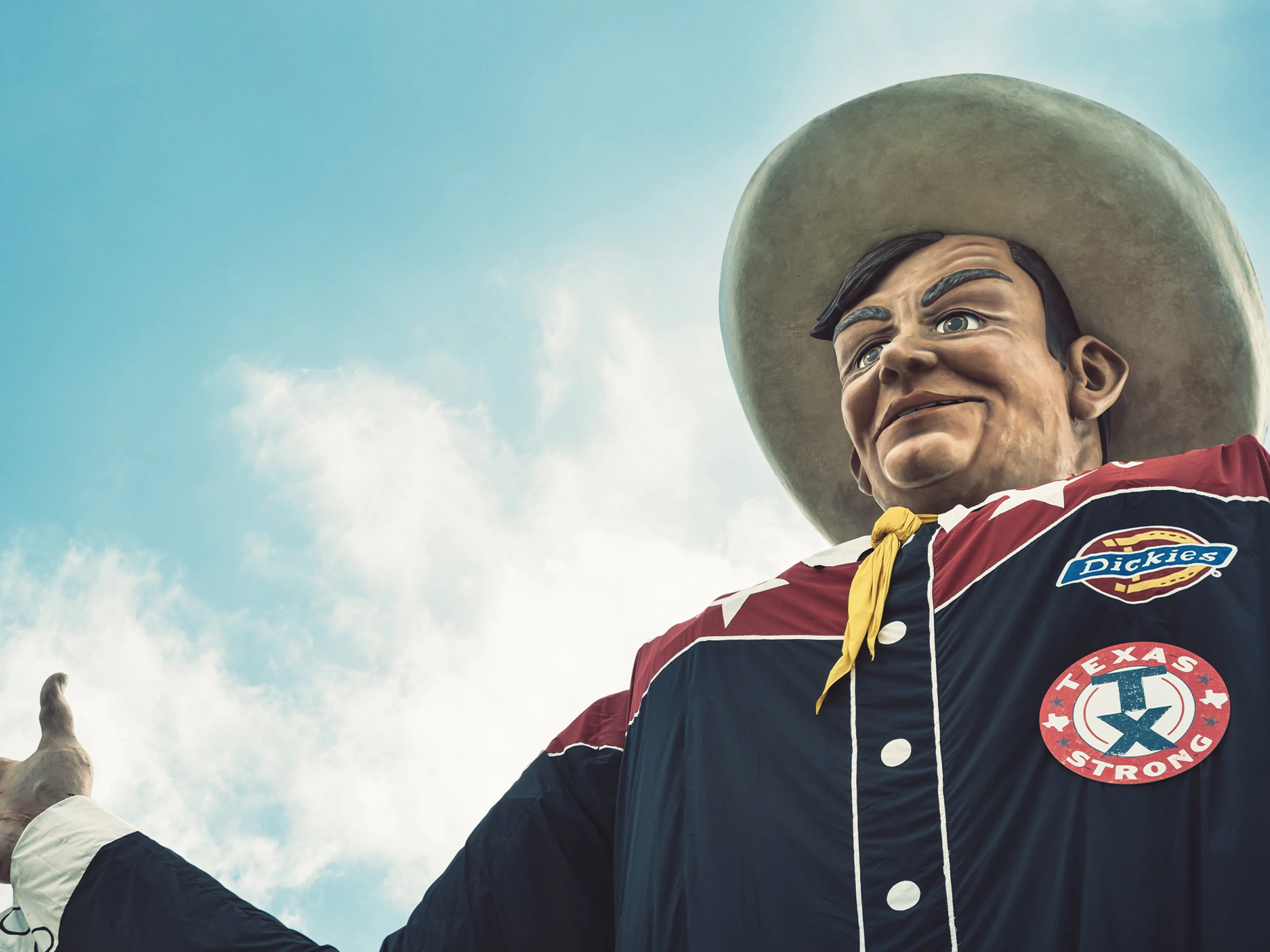 A large cowboy statue in a wide-brimmed hat and dark shirt stands with arms open against a blue sky with clouds. The shirt features patches, including one reading TEXAS STRONG.