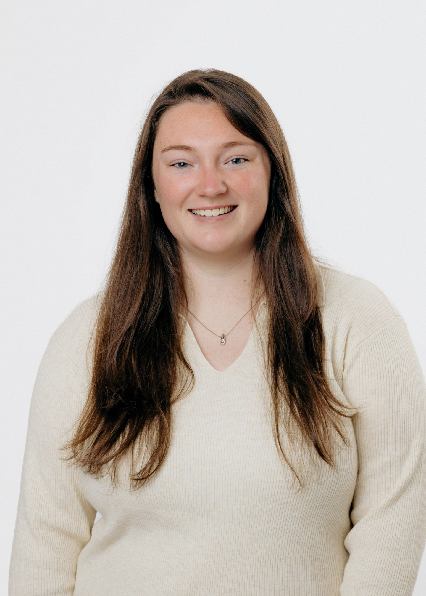 A woman with long brown hair wearing a cream-colored sweater smiles at the camera against a plain white background.