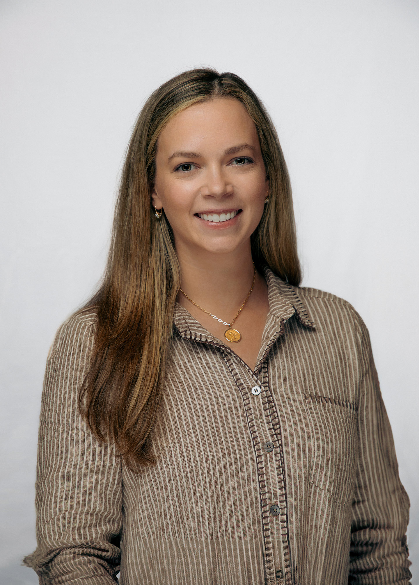 A woman with long brown hair, wearing a brown and white striped button-up shirt and gold necklaces, smiles at the camera against a plain light background.