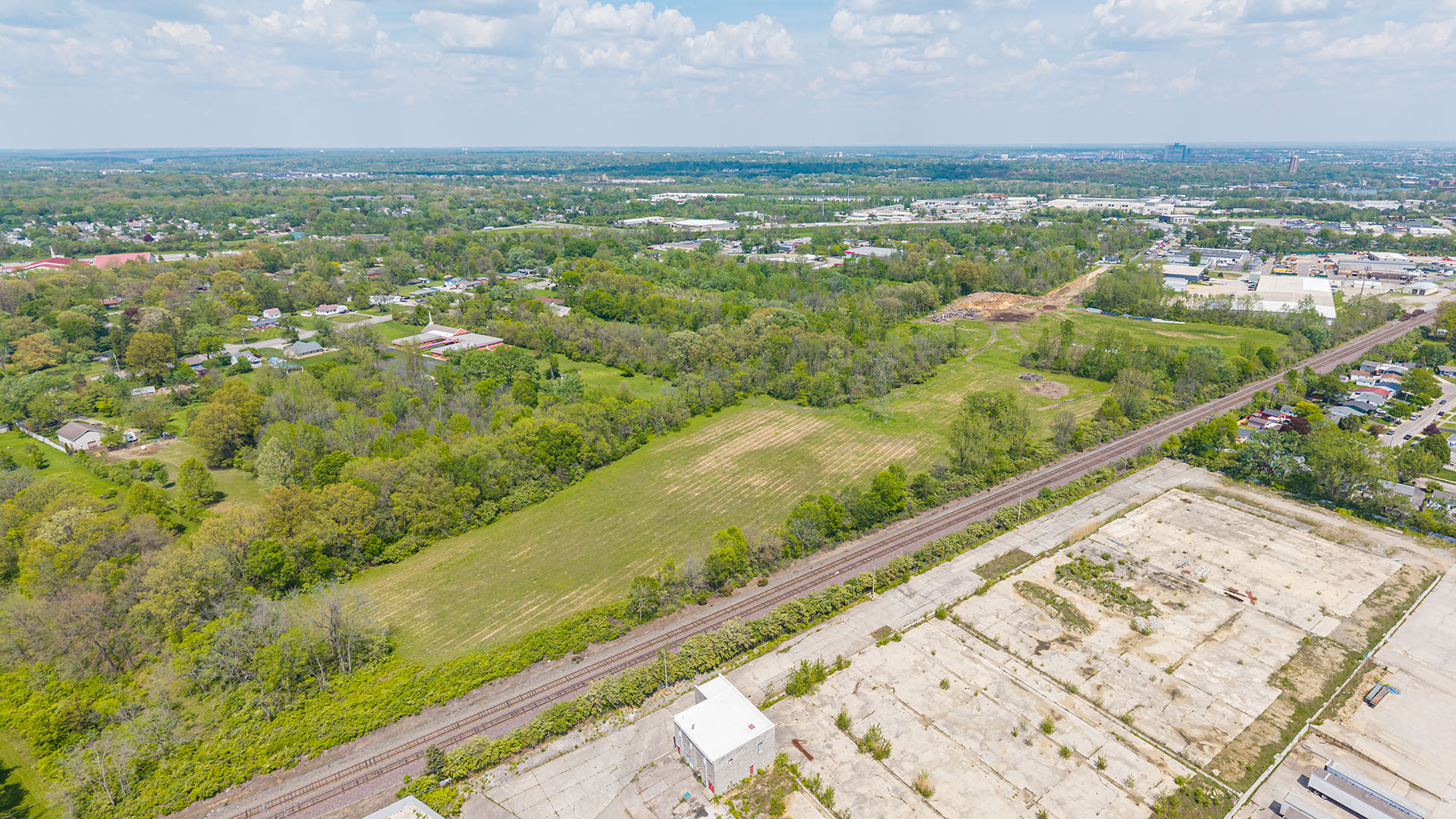 Aerial view of a green, tree-lined rural area with open grassy fields, a railway track, and an adjacent paved industrial site under a partly cloudy sky.