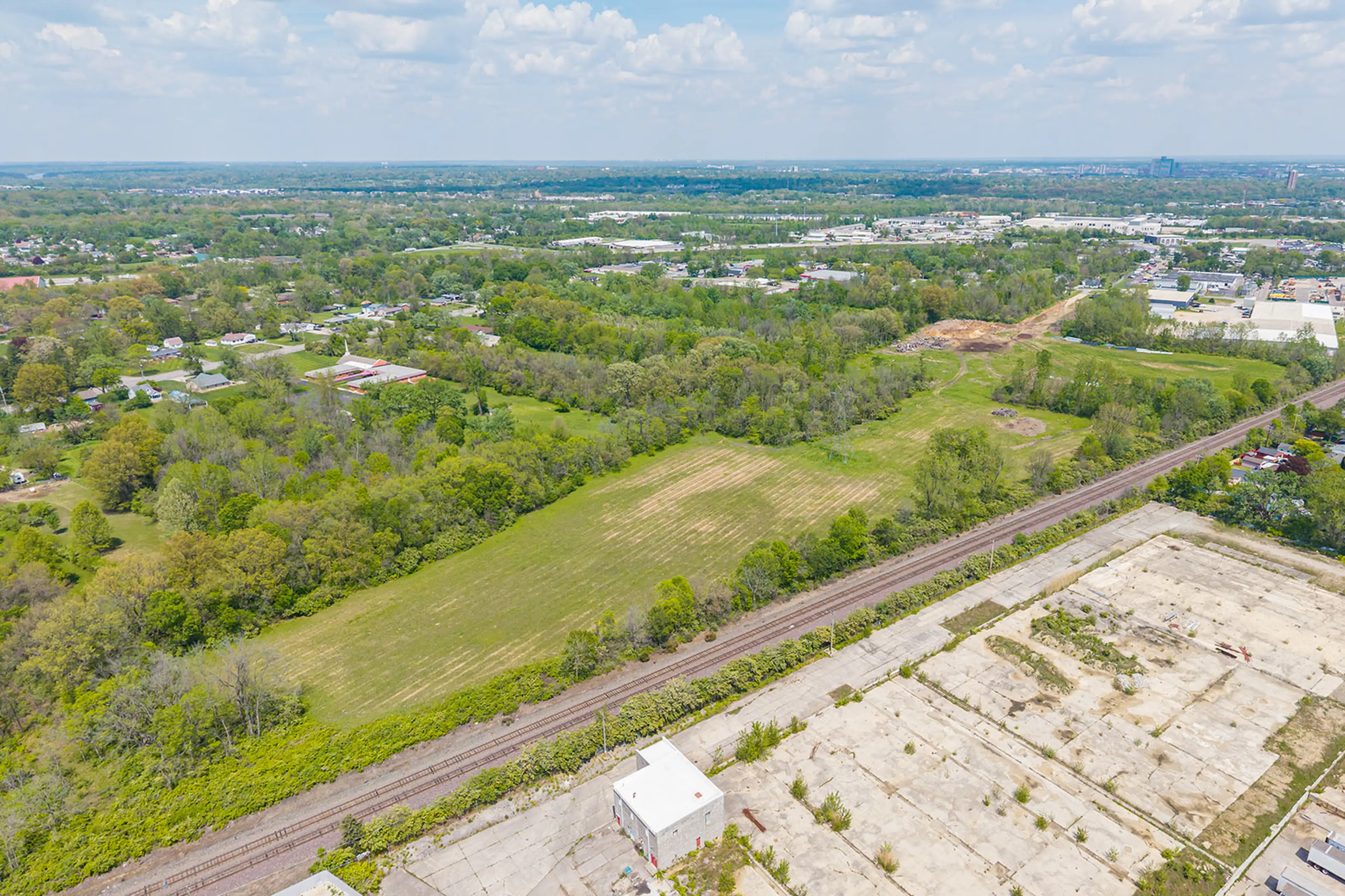 Aerial view of a green, tree-lined rural area with open grassy fields, a railway track, and an adjacent paved industrial site under a partly cloudy sky.
