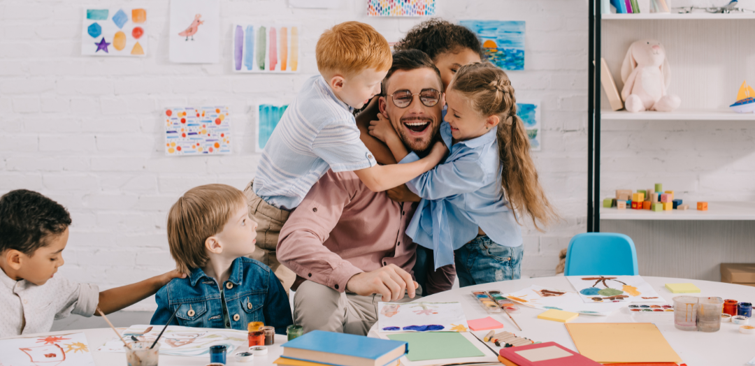 Young students hugging a male teacher sitting at a table in a classroom