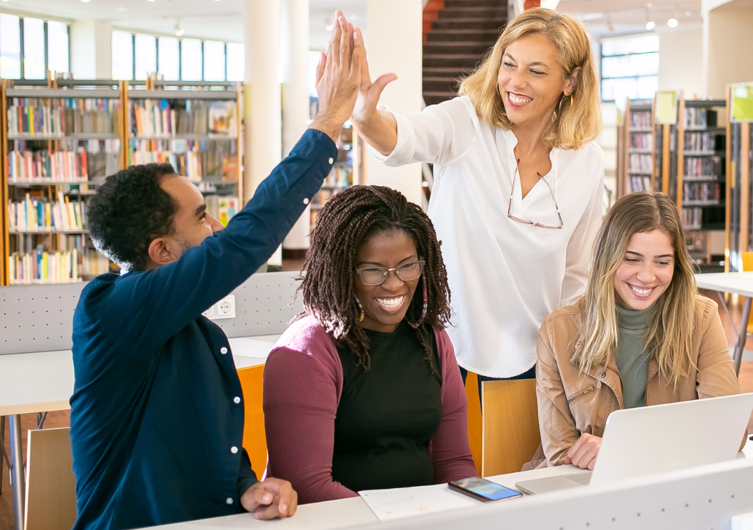 A mentor teacher is high-fiving a male student teacher with two female student teachers sitting with them in the school library.