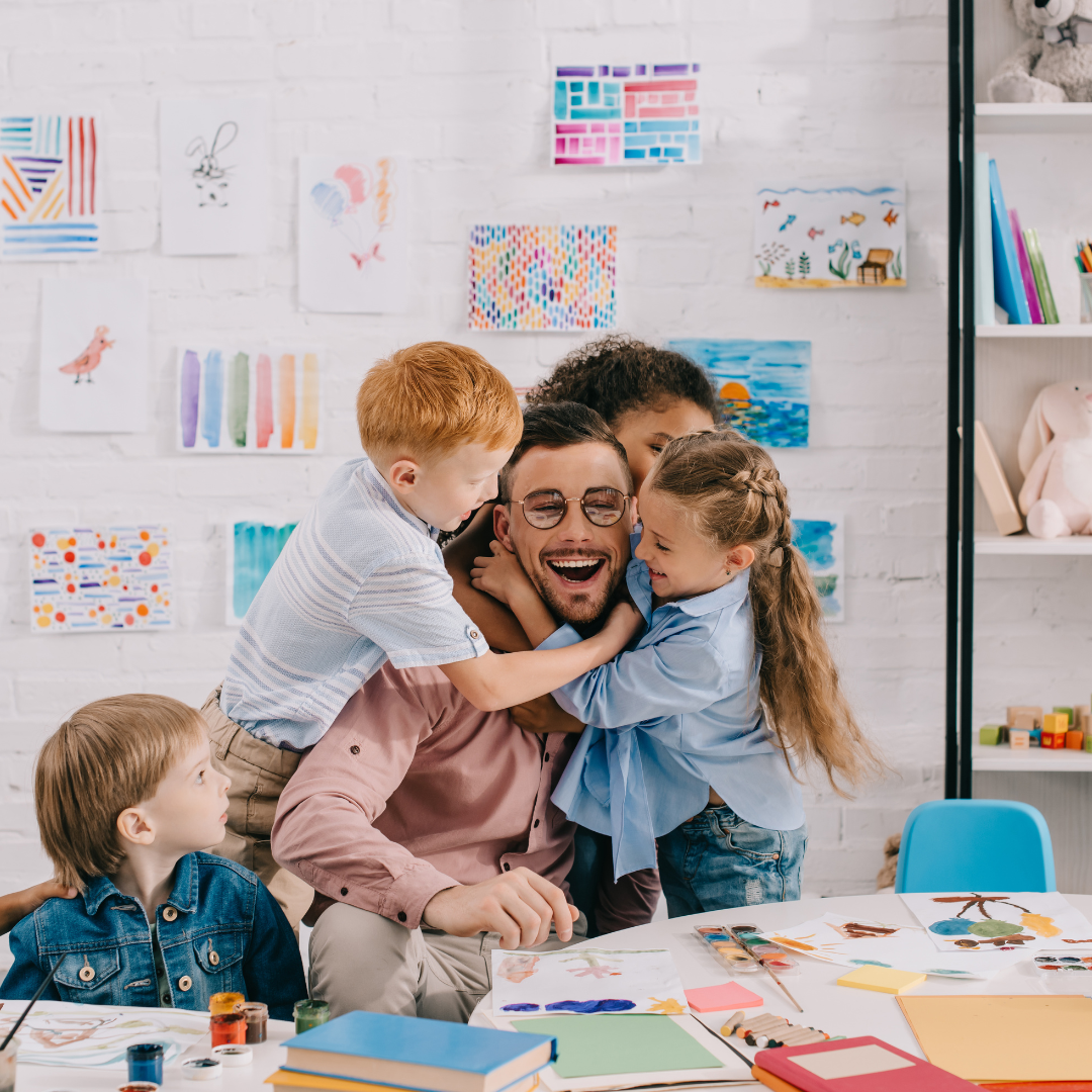 Young students hugging a male teacher sitting at a table in a classroom