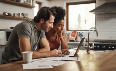 Two people sitting at a table looking at a laptop with paper around it.