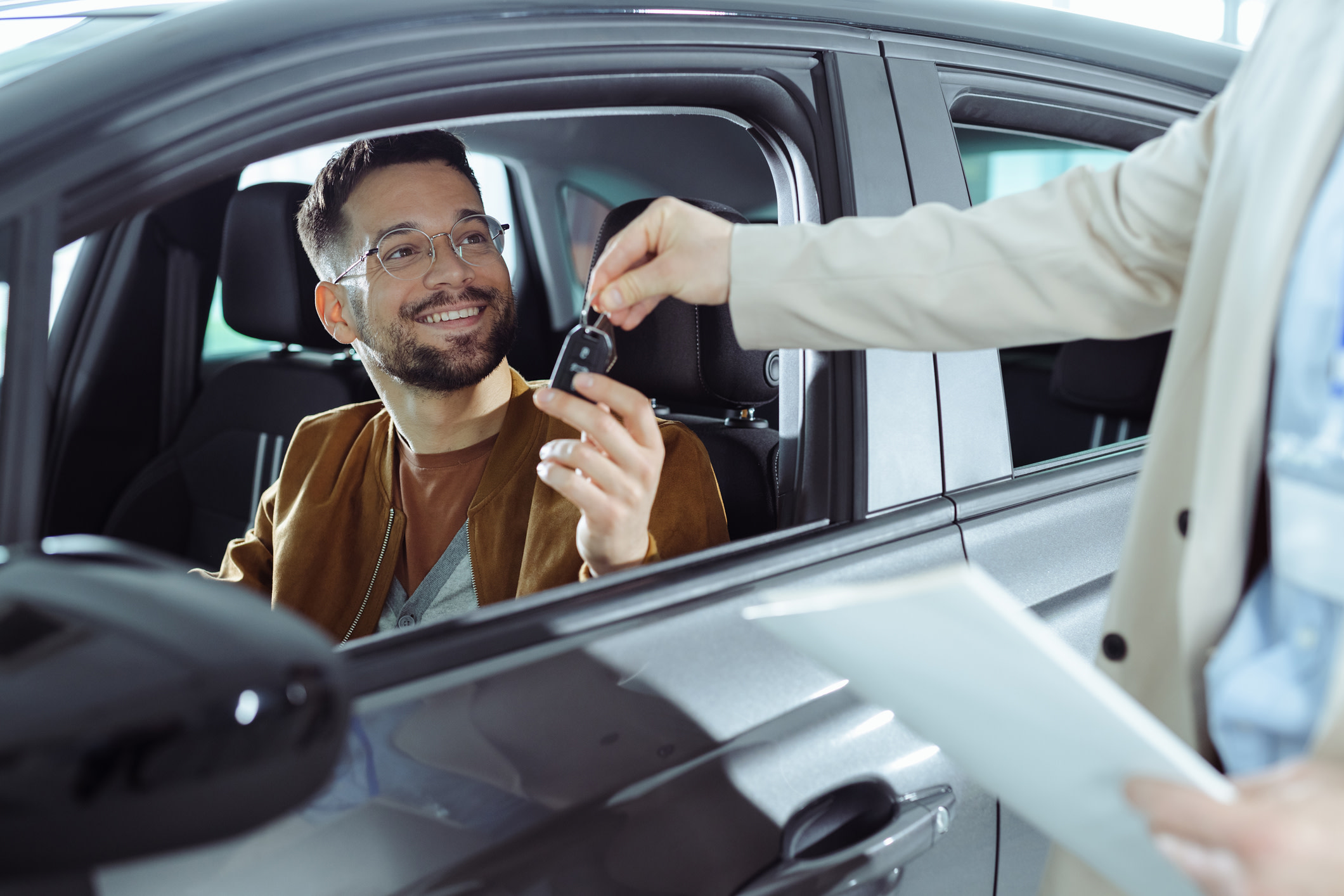 A man with glasses and a brown jacket in a car being handed keys to a rental vehicle. 