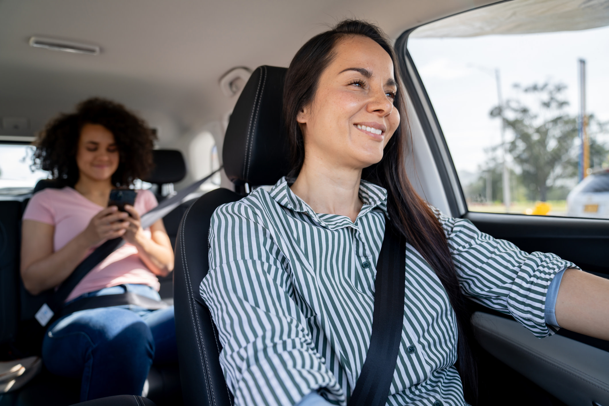 A woman driving with another woman in the backseat on her phone.