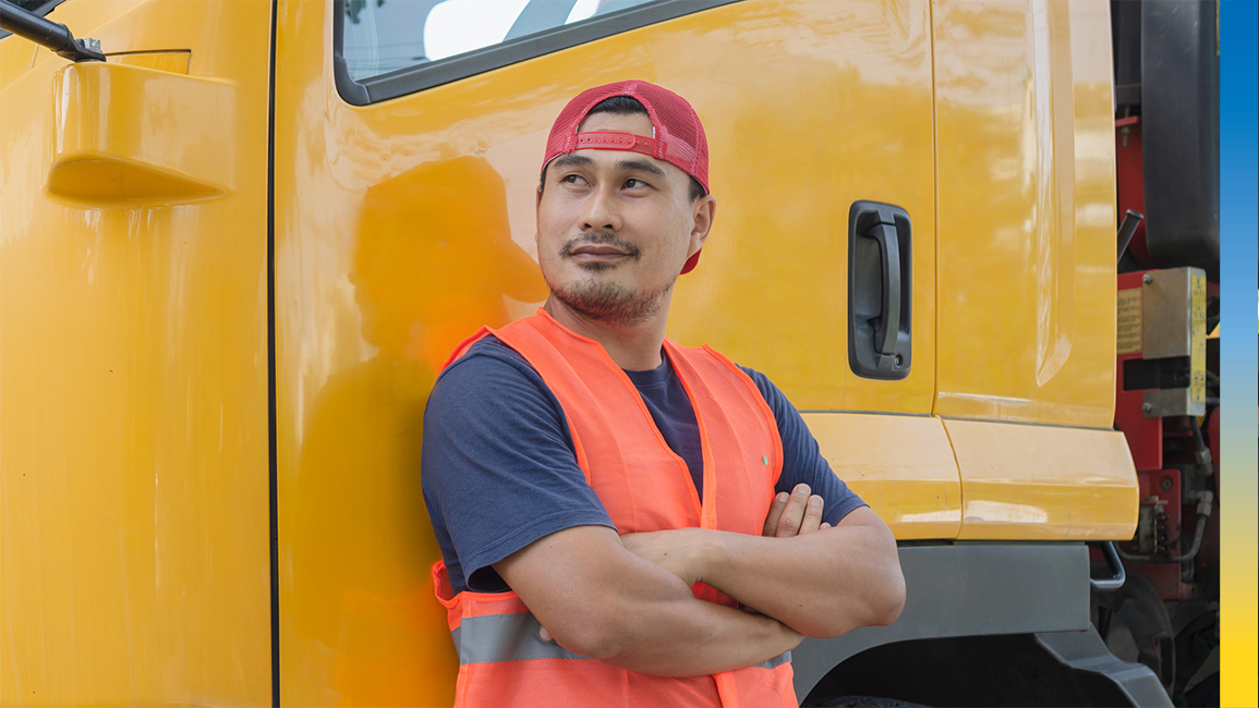 A man in a red hat and an orange safety vest leaning against the side of a yellow truck.