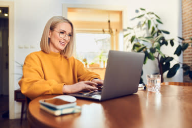 A woman wearing a yellow shirt sitting at a wooden table looking at a silver laptop.