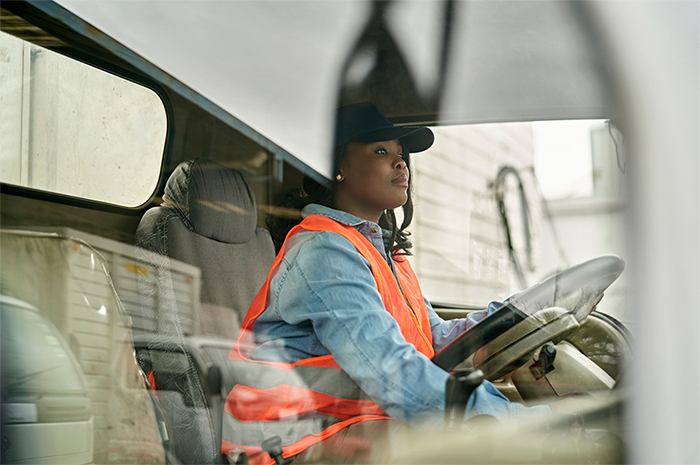 A woman inside the cab of a truck wearing an orange safety vest and a black baseball cap