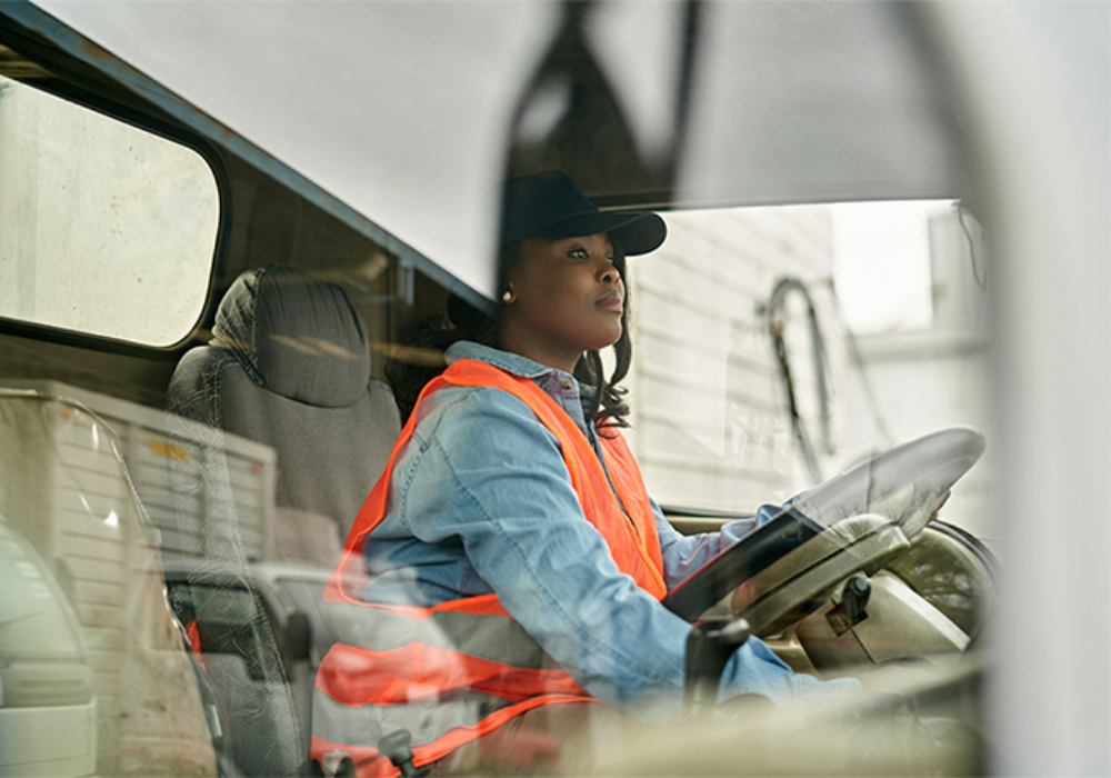 A woman inside the cab of a truck wearing an orange safety vest and a black baseball cap