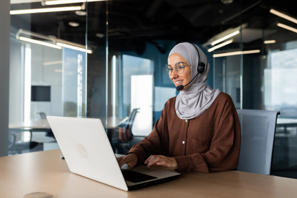 A young woman wearing a hijab and a headset is working in the office at a desk using a laptop.