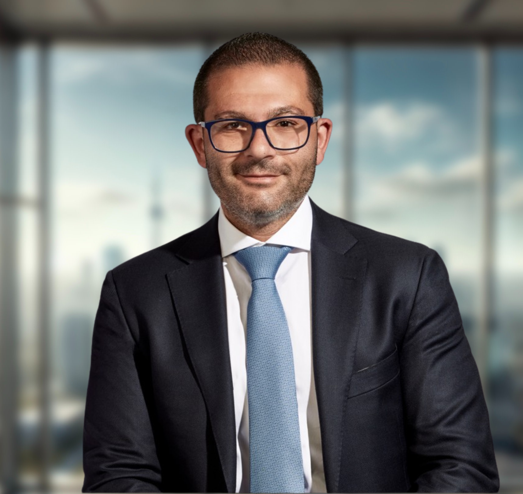 A man with short dark hair and a short beard, wearing glasses, and a dark blazer with a white shirt and light blue tie.