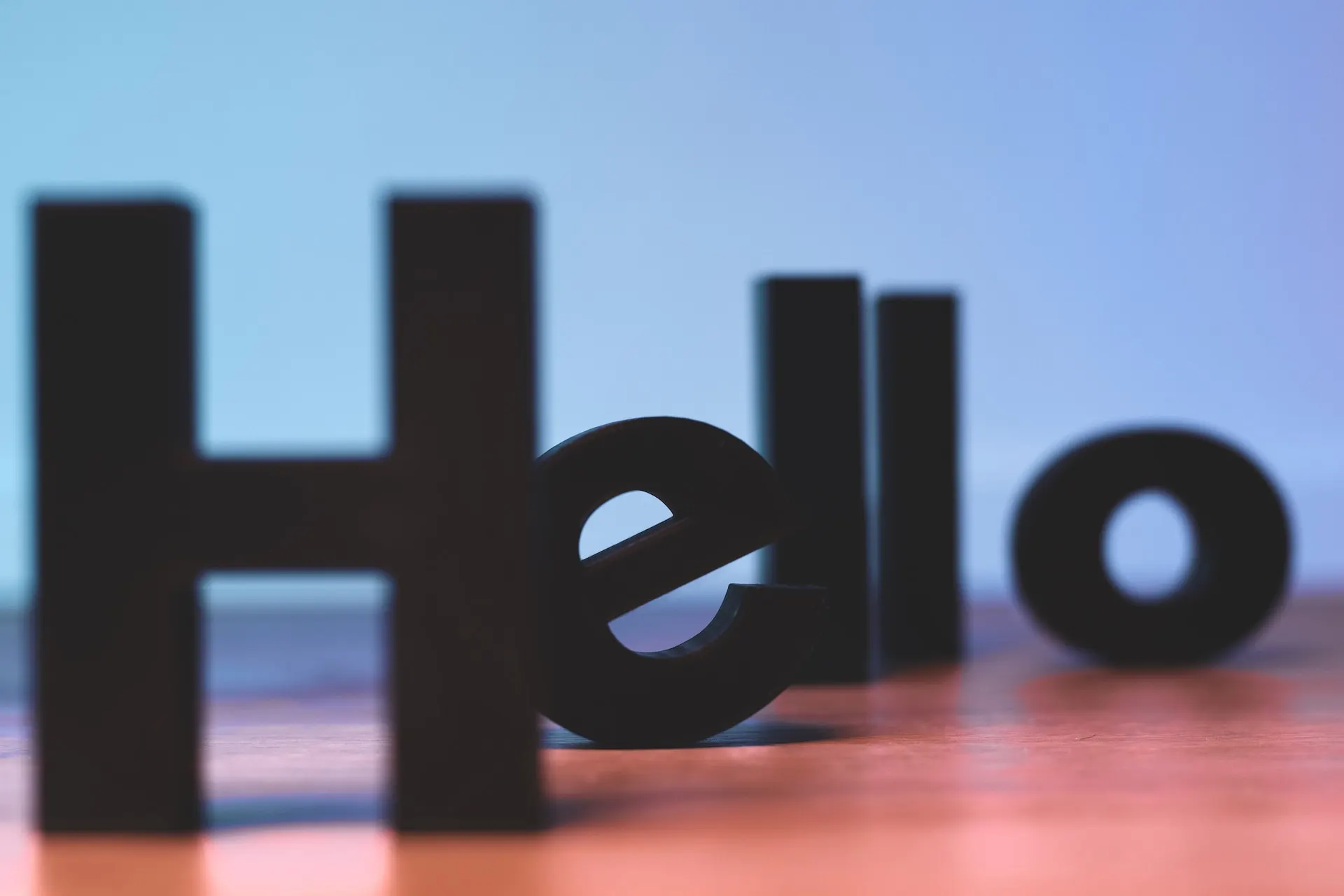 Vemos a palavra Hello feita com letras que são objetos pretos em cima de superfície de madeira.
